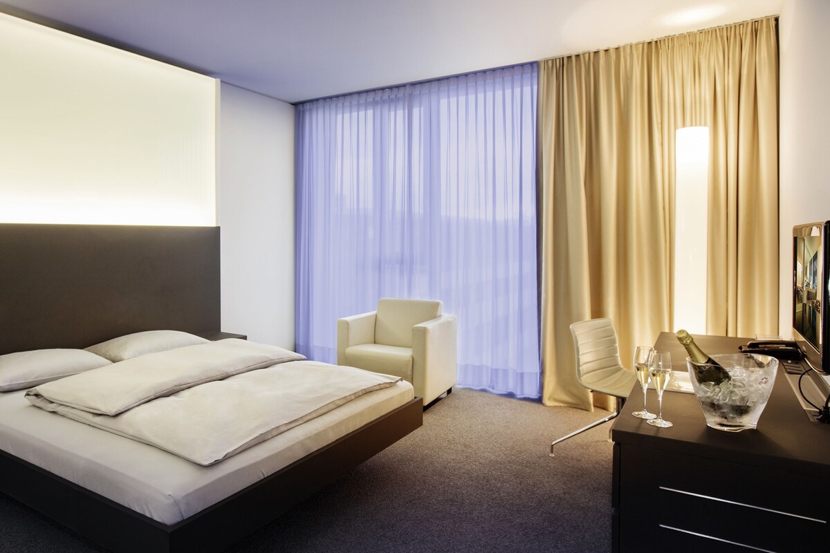 A hotel room with a bed, two chairs, a desk with a TV, and a minibar.