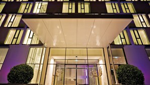 A modern hotel entrance with a glass facade, illuminated windows, and a central entrance area with a reception desk. - INNSiDE by Meliá Düsseldorf Derendorf (Düsseldorf)
