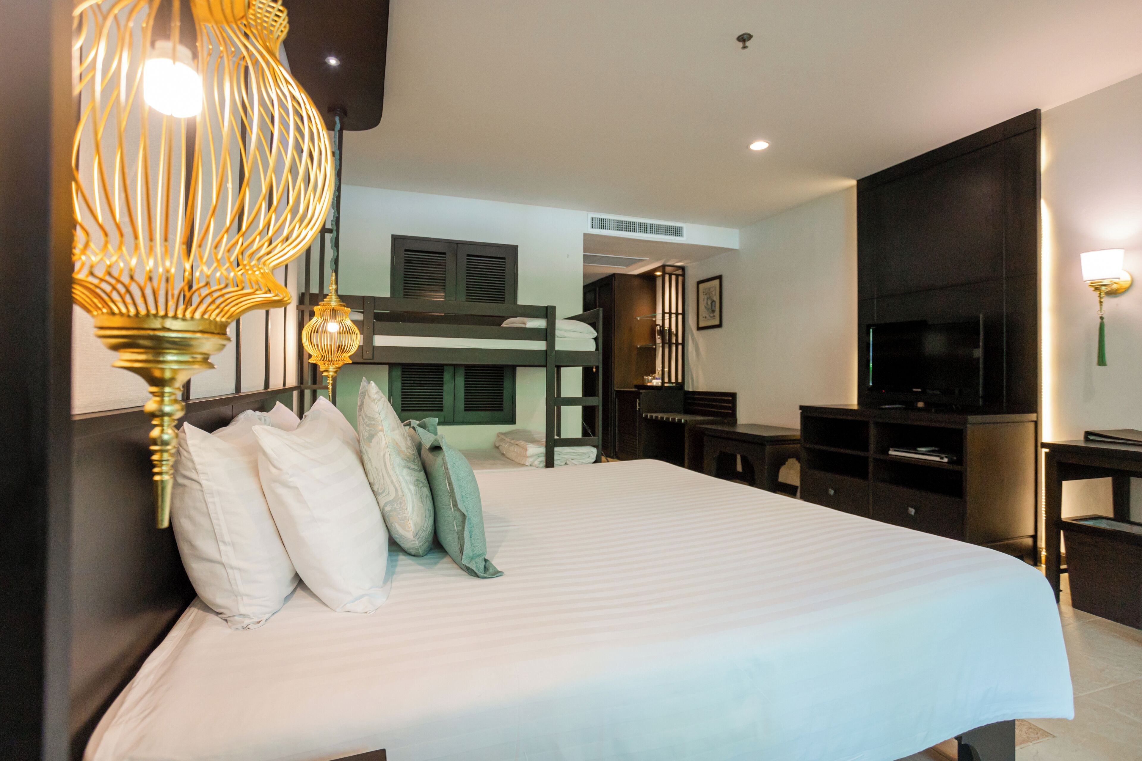 A hotel room with a large bed, a TV, a desk, and a bunk bed in the background.