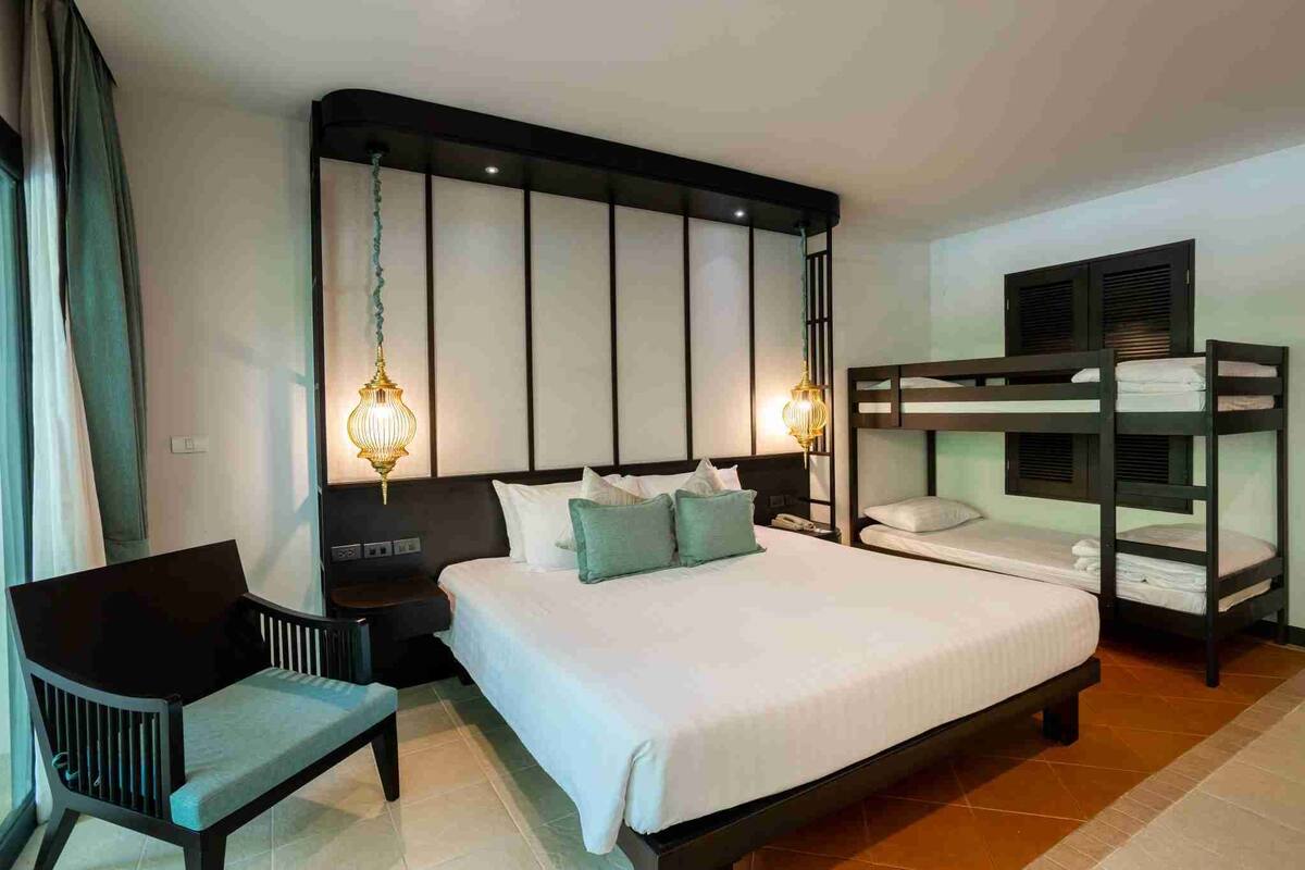 Quarto de hotel com uma cama grande, duas mesas de cabeceira, uma cadeira, uma cama beliche e duas luminárias fixadas na parede.