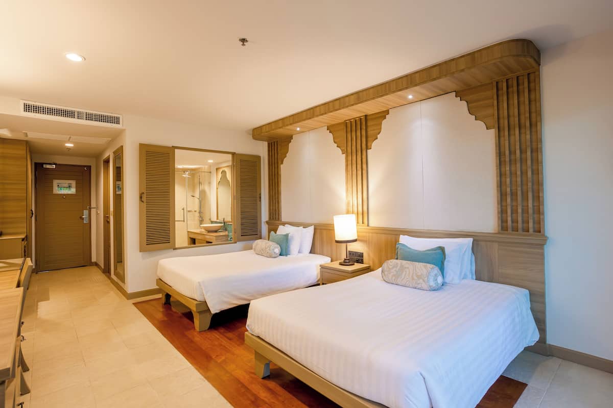 Quarto de hotel com duas camas, cabeceira de madeira, mesa de cabeceira com abajur, espelho e porta de madeira.
