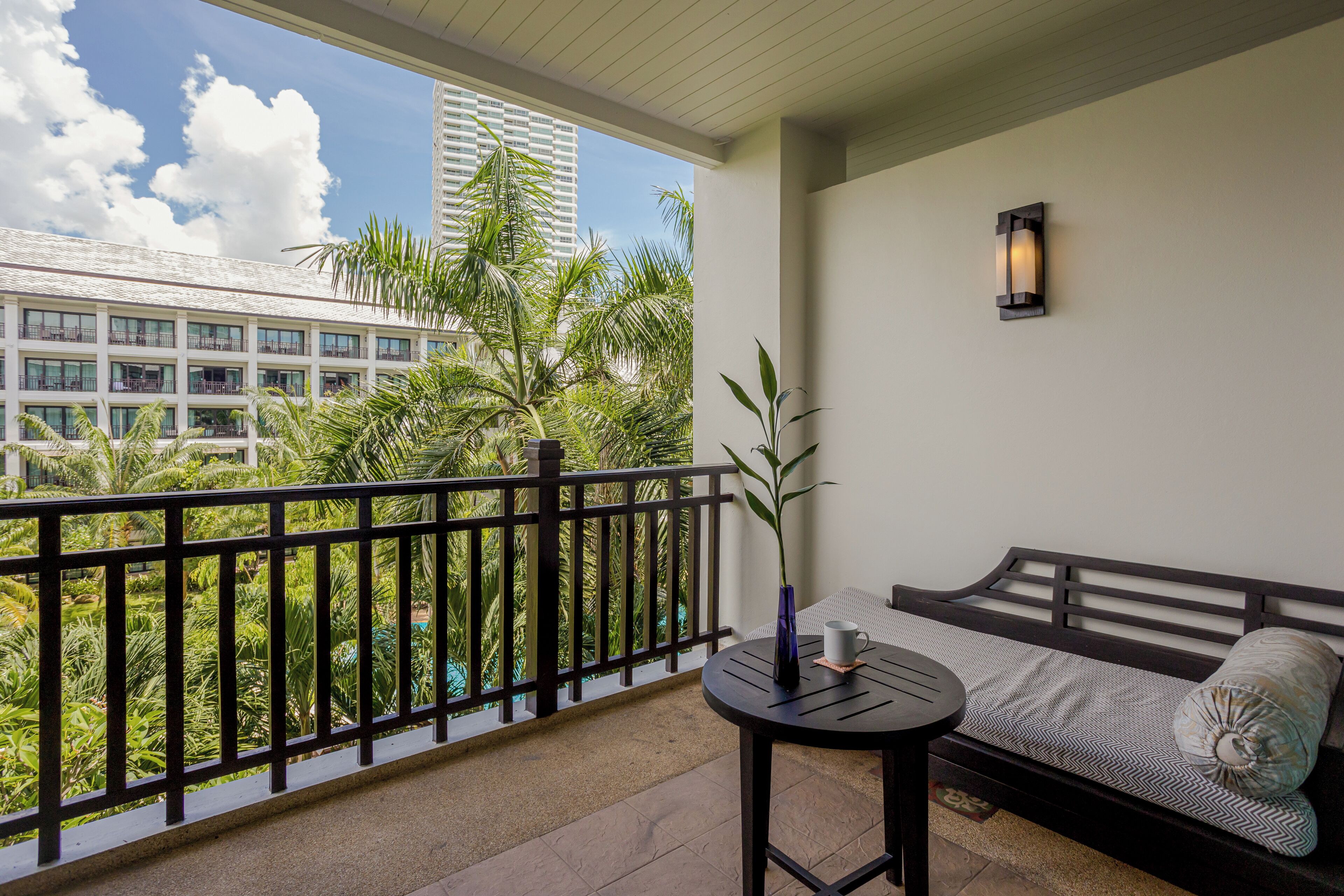 A balcony with a view of a building and palm trees, a small round table with a vase and a cup, and a sofa with patterned cushions.