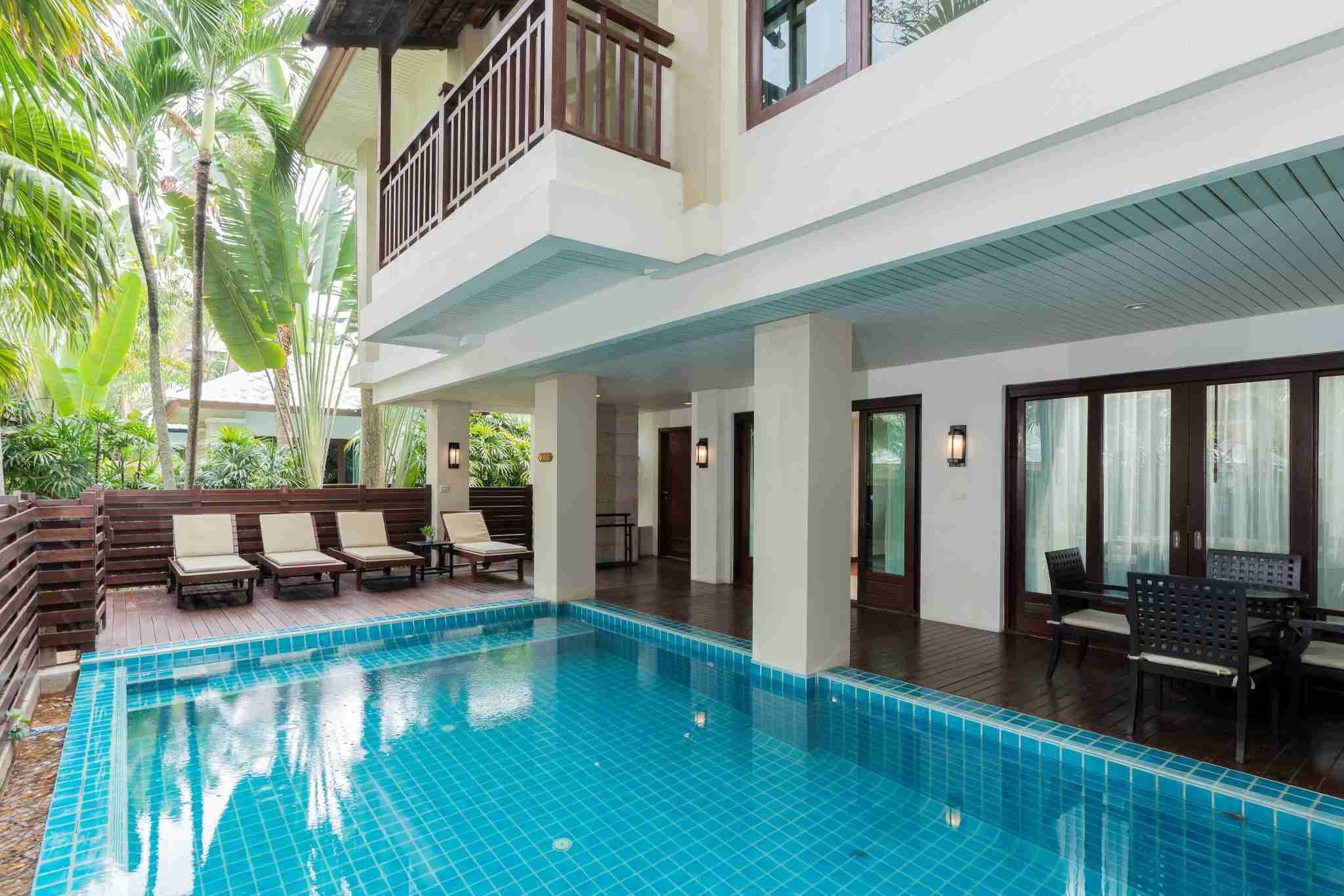 A swimming pool with lounge chairs and a covered area, surrounded by palm trees and a two-story building with balconies.