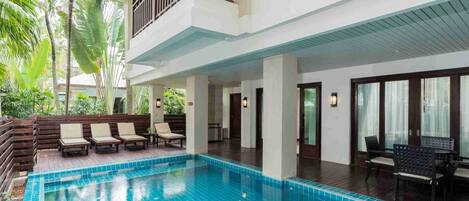 A swimming pool with lounge chairs and a covered area, surrounded by palm trees and a two-story building with balconies.