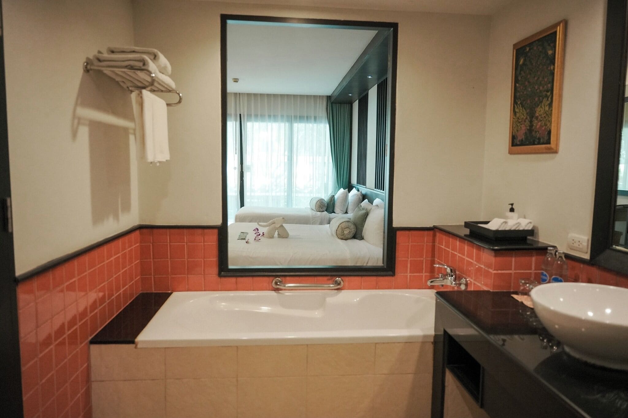 Une salle de bain moderne avec une grande baignoire, un lavabo et un miroir qui reflète une chambre à coucher avec un lit et des rideaux.