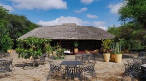 An outdoor dining area with thatched-roof structures, surrounded by potted plants and metal chairs.