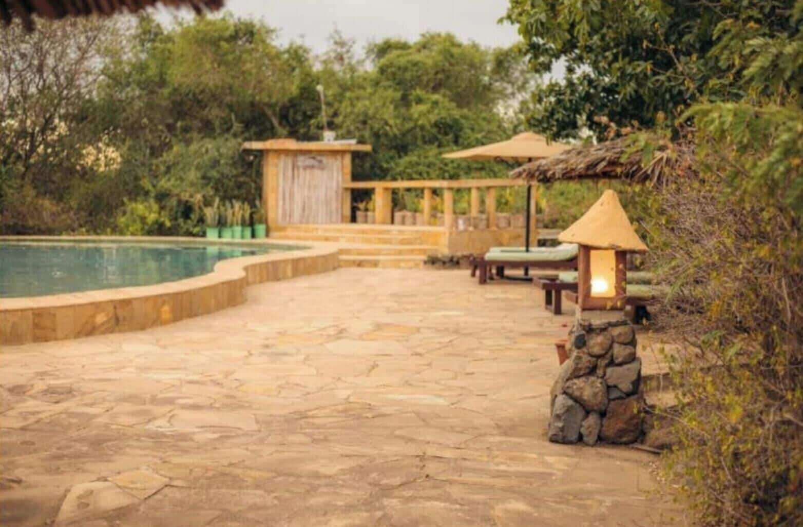 A stone patio leading to a swimming pool, surrounded by lush greenery and a wooden structure with a thatched roof.