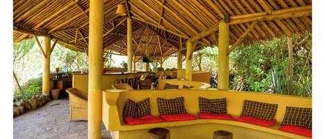 A covered outdoor seating area with a thatched roof, yellow walls, and red cushions.
