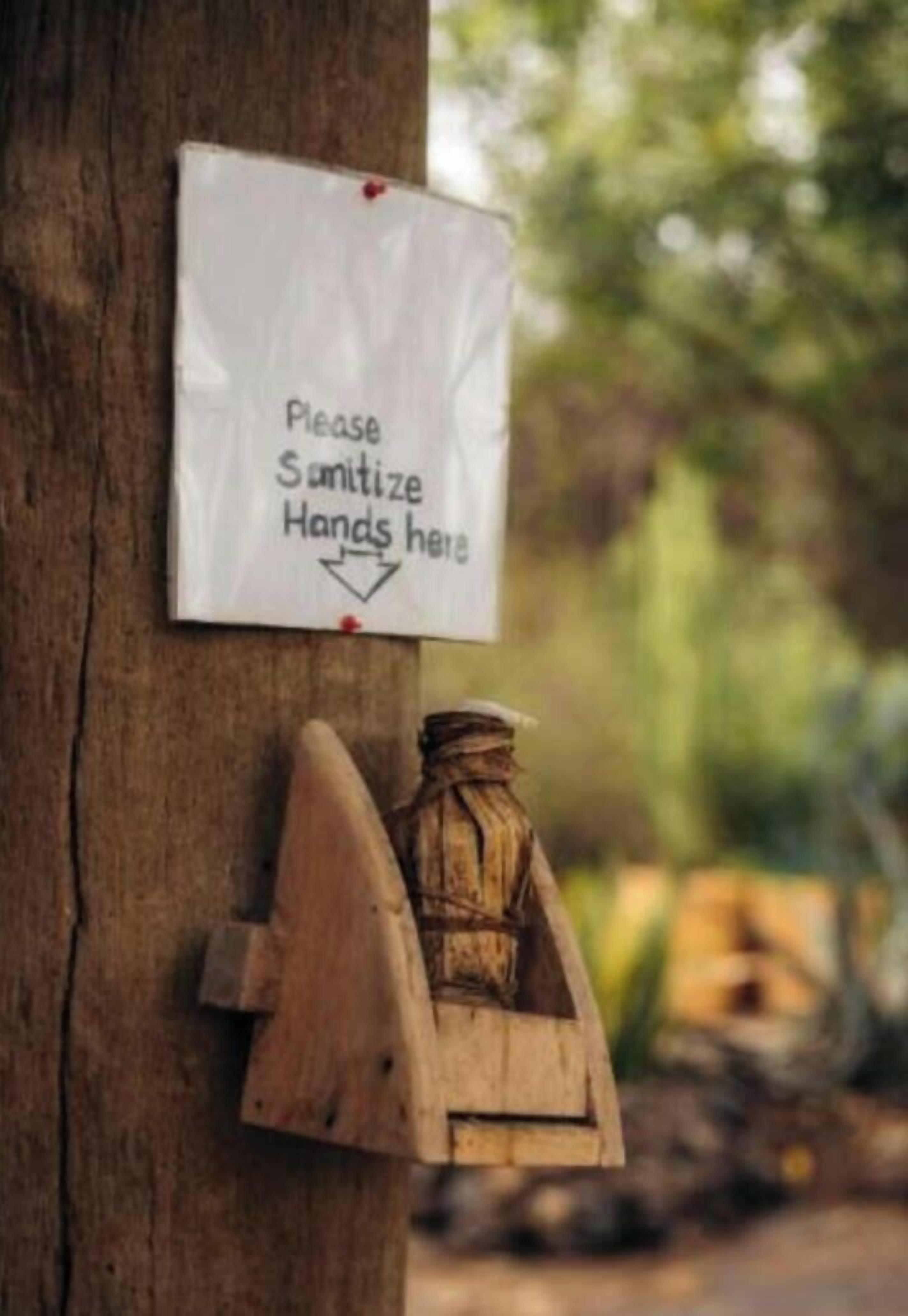 Schild an einem Baum, das um Handdesinfektion bittet, mit einem Holz-Händedesinfektionsspender darunter.