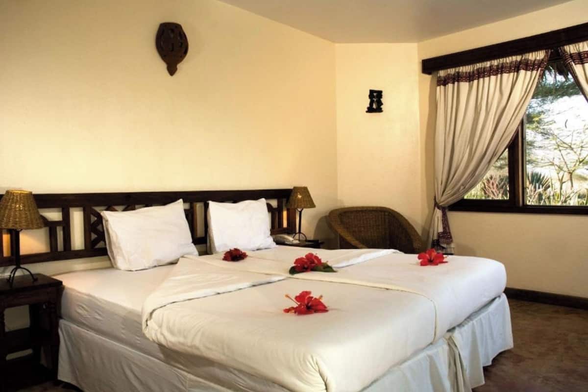 Una cama bien hecha con sábanas blancas y flores rojas, cabecera de madera, dos lámparas de noche, una mesita de noche de madera y una ventana con cortinas.