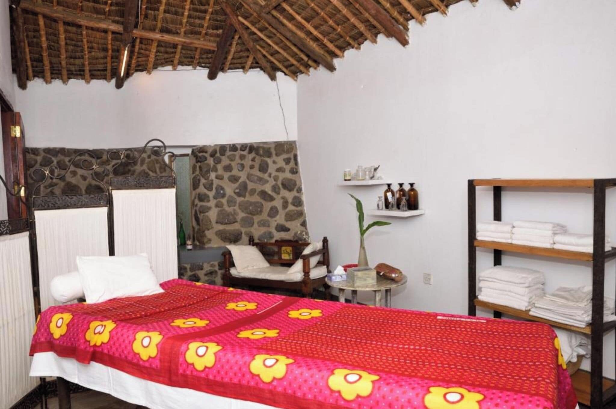 A room with a bed, a chair, a small table, a shelf with toiletries, and a wooden shelf with folded towels.