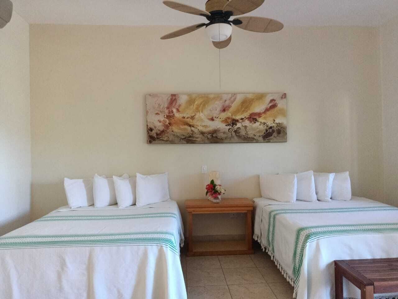 Una stanza con due letti, una ventola a soffitto, un quadro appeso al muro e un tavolino con un vaso di fiori.
