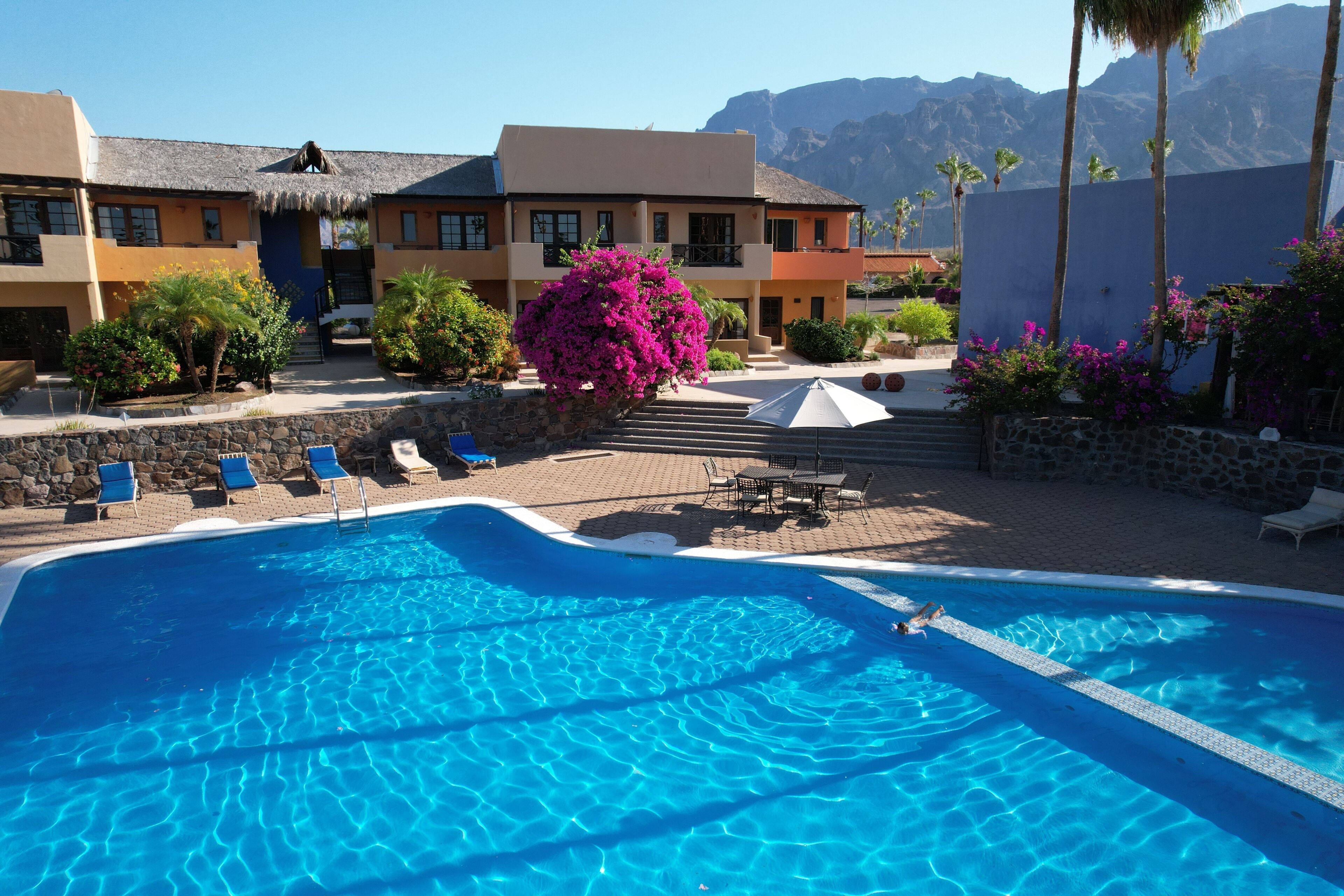 A swimming pool with lounge chairs, a thatched-roof building, and a mountain backdrop.