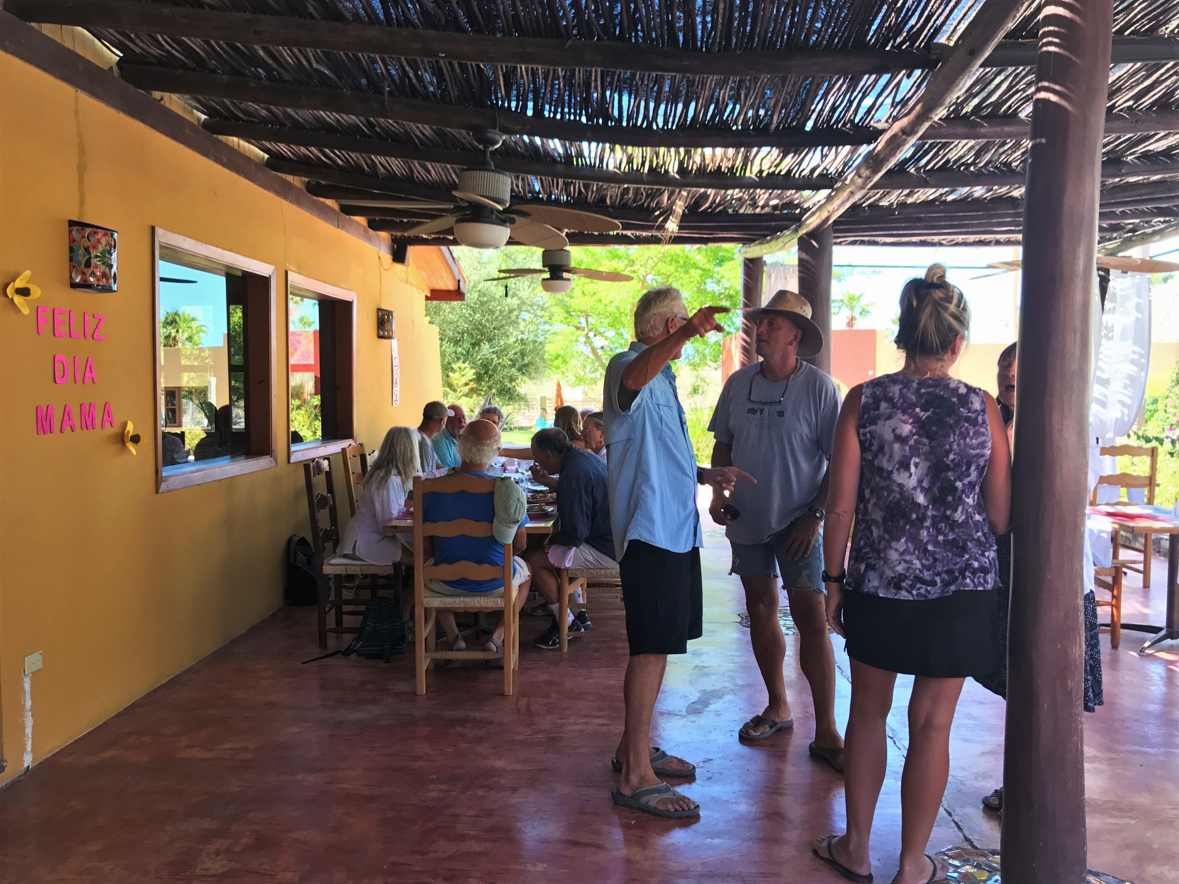 A covered outdoor dining area with wooden tables and chairs, a thatched roof, and a wall with a 'Feliz Dia Mama' sign.