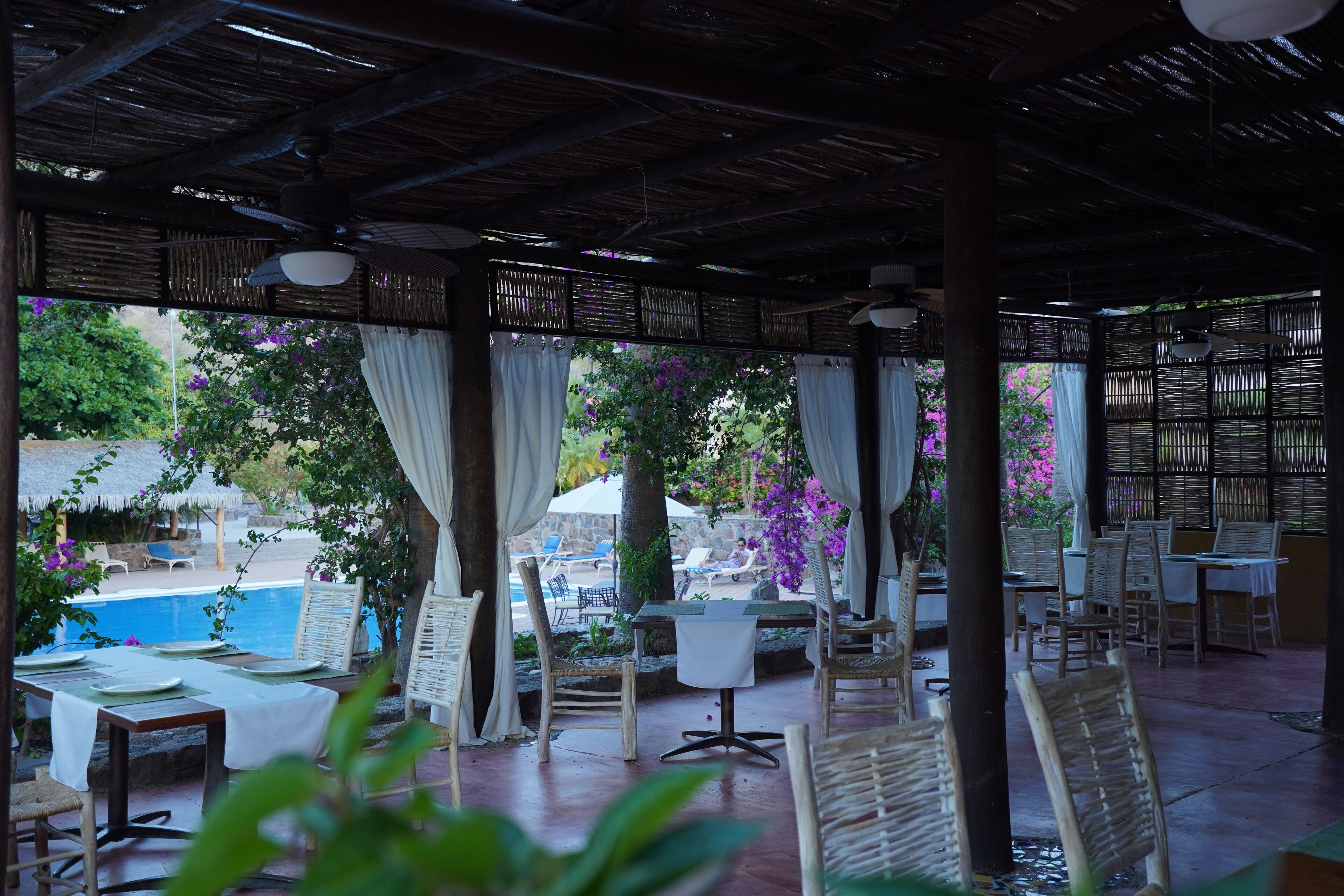 An outdoor patio area with tables and chairs, a pool in the background, and a view of the surrounding landscape.
