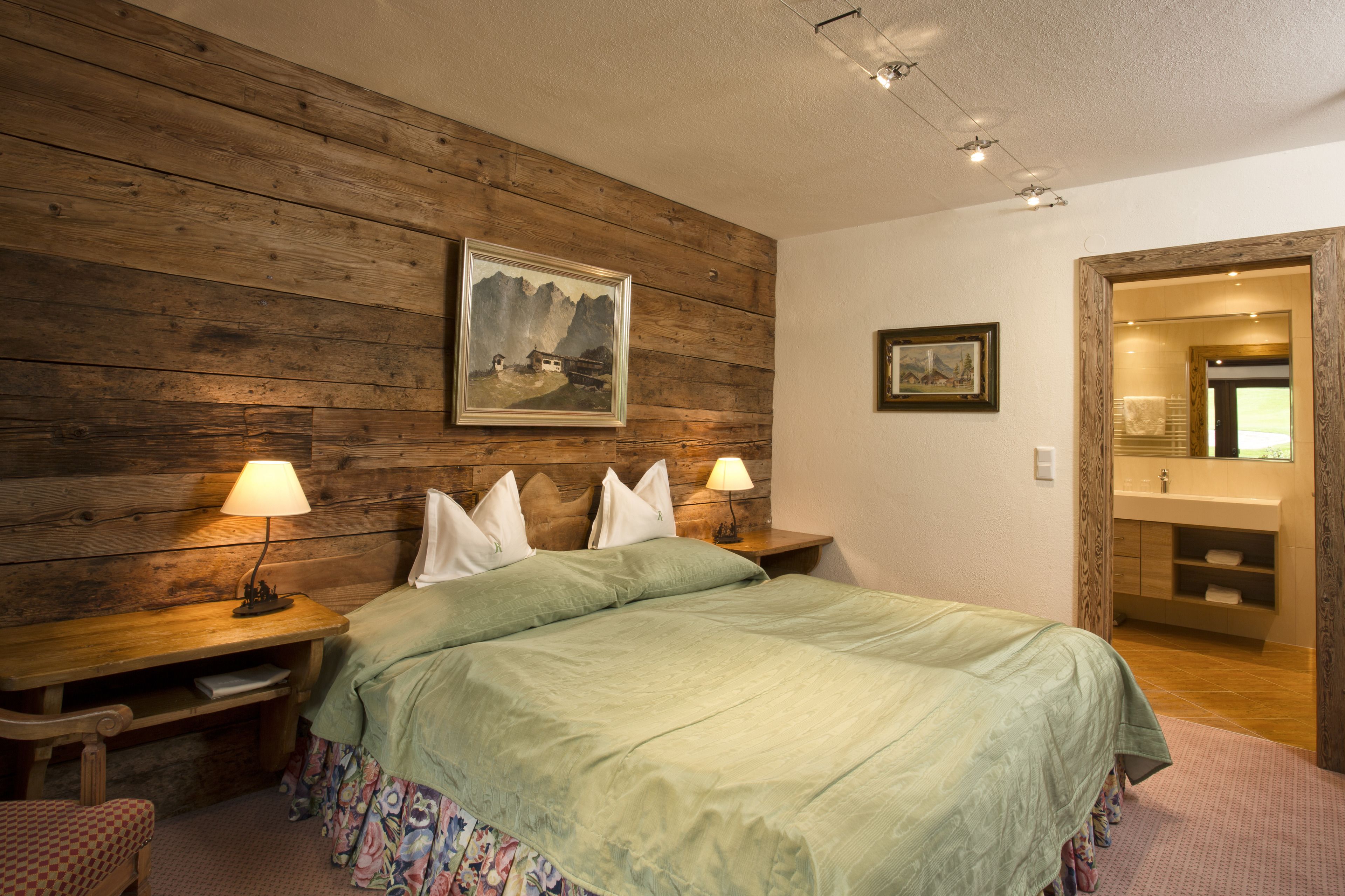 A bedroom with a wooden headboard, two bedside tables with lamps, a bed with a green bedspread, and a bathroom visible through an open door.