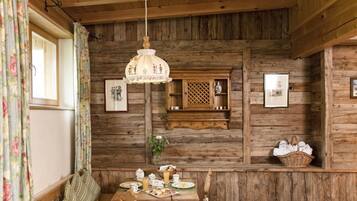 A rustic wooden cabin interior with a dining table set for breakfast, a wooden bench, and a hanging light fixture.