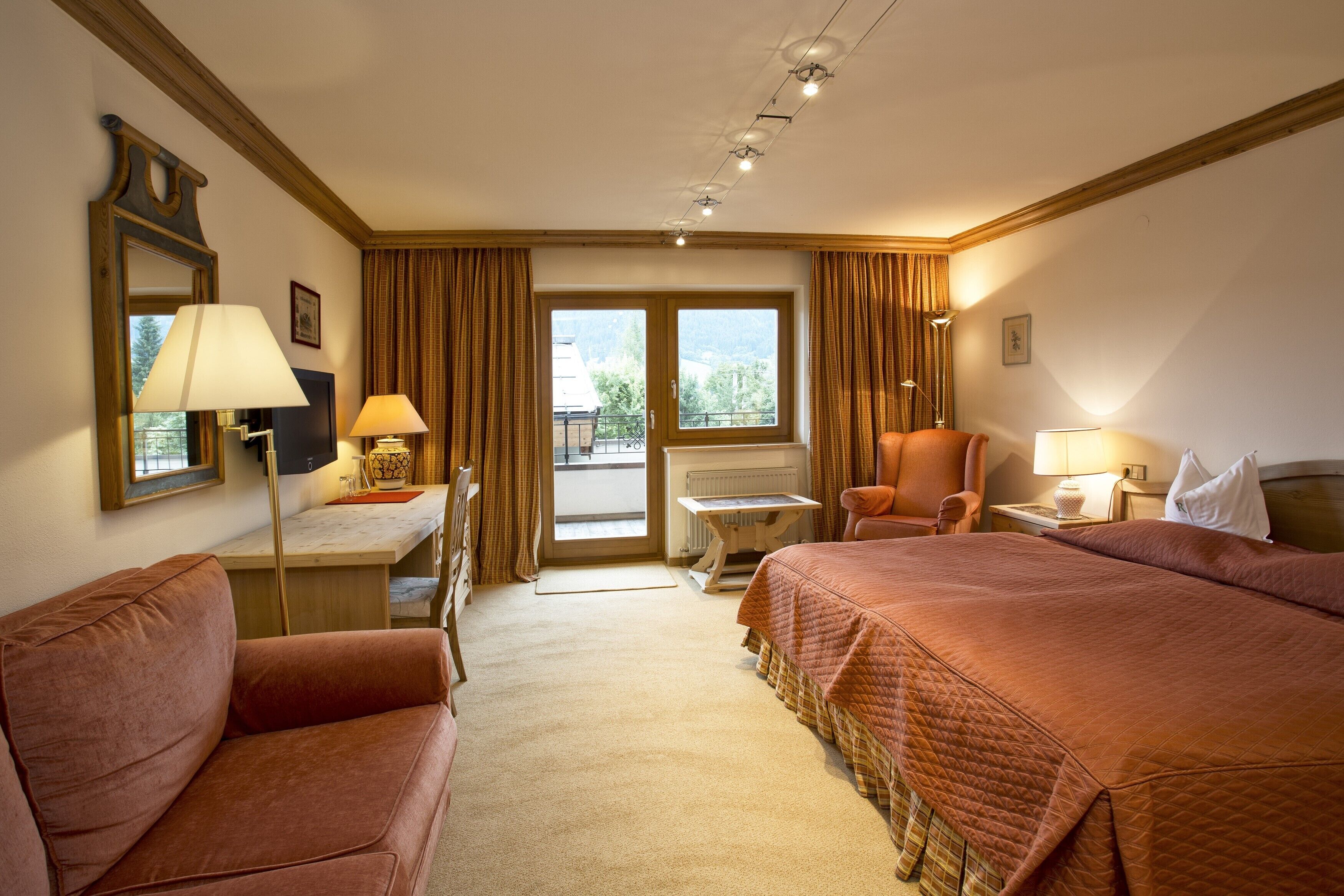 Habitación de hotel con dos camas, un sofá, un escritorio y una silla. Tiene una ventana con cortinas, un radiador y una lámpara sobre el escritorio.