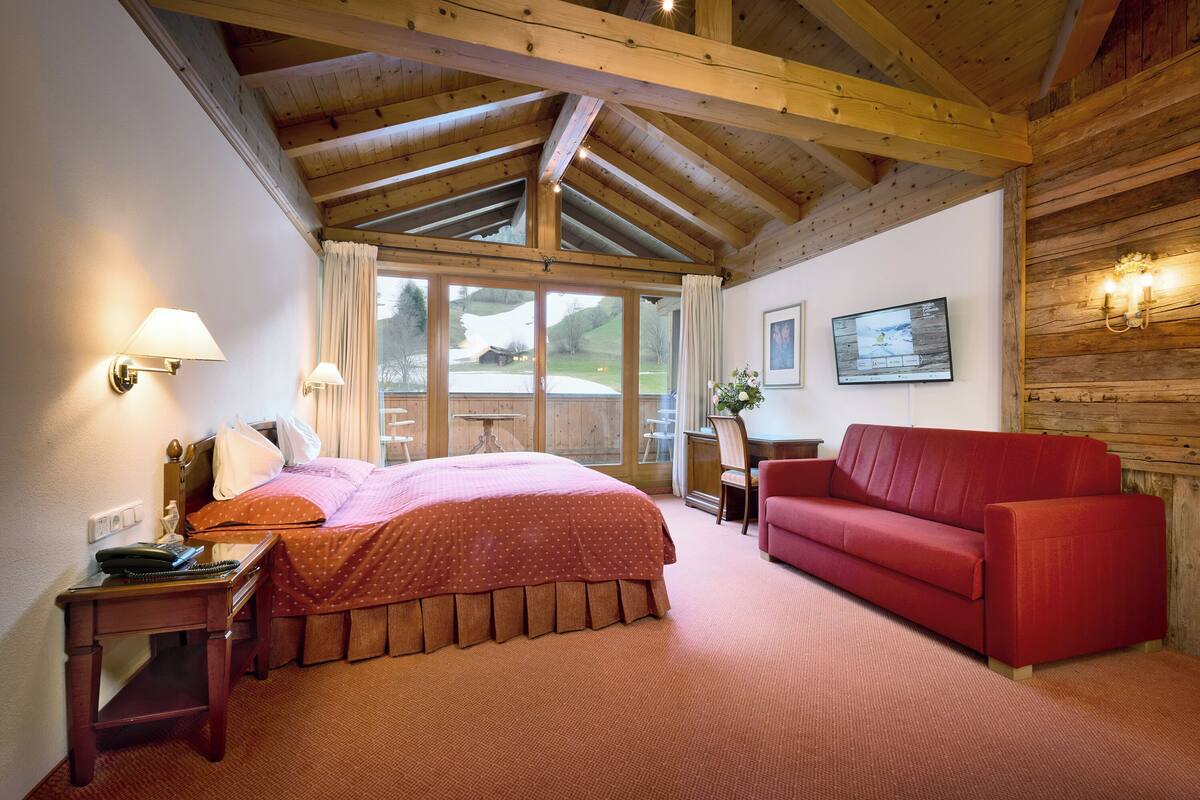 Une chambre d’hôtel avec un plafond en bois, un lit recouvert d’une couvre-lit à pois, un canapé rouge, un bureau avec une lampe et un téléviseur fixé au mur.