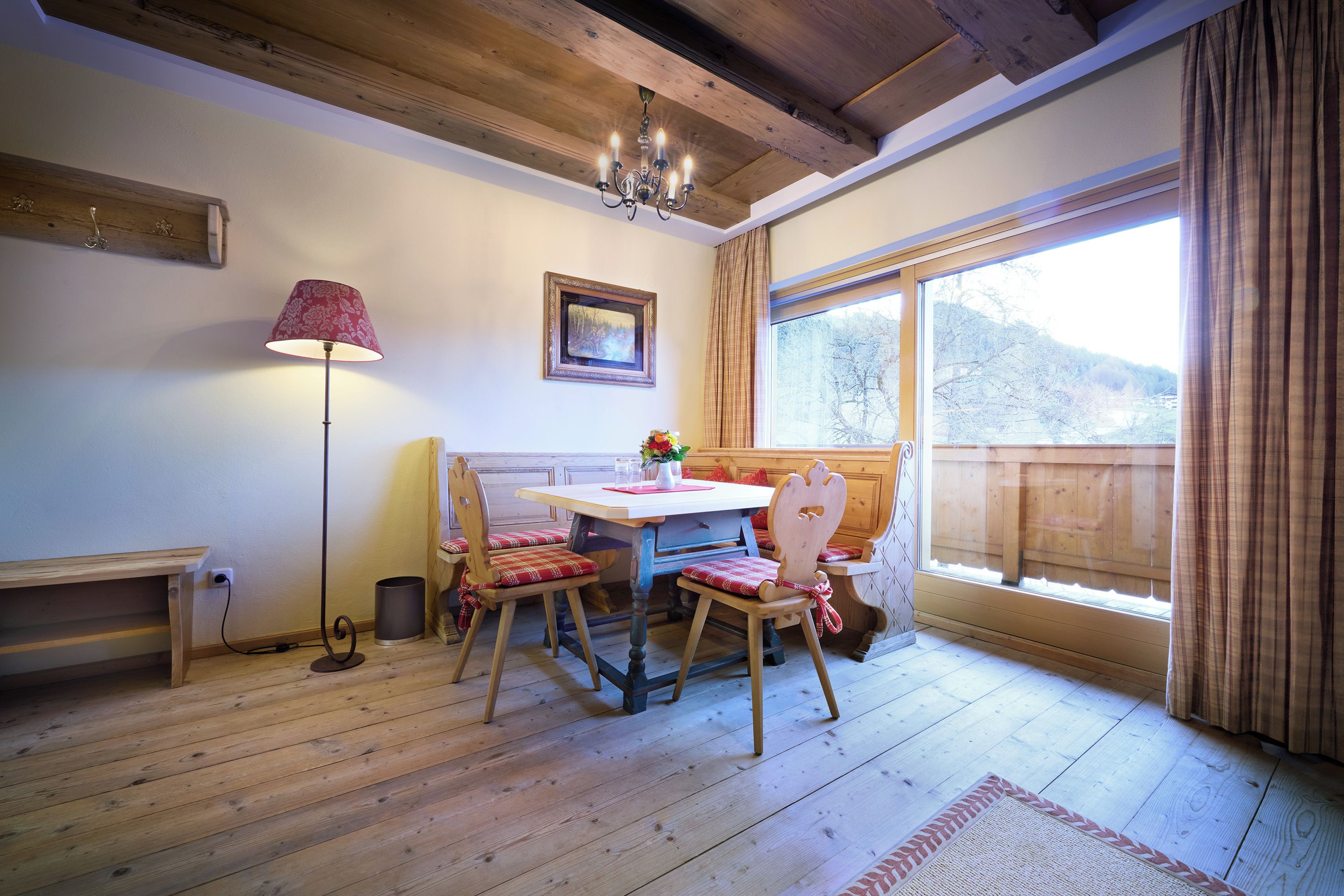 Un comedor con mesa y sillas de madera, una araña, un cuadro y un ventanal con vista a árboles.