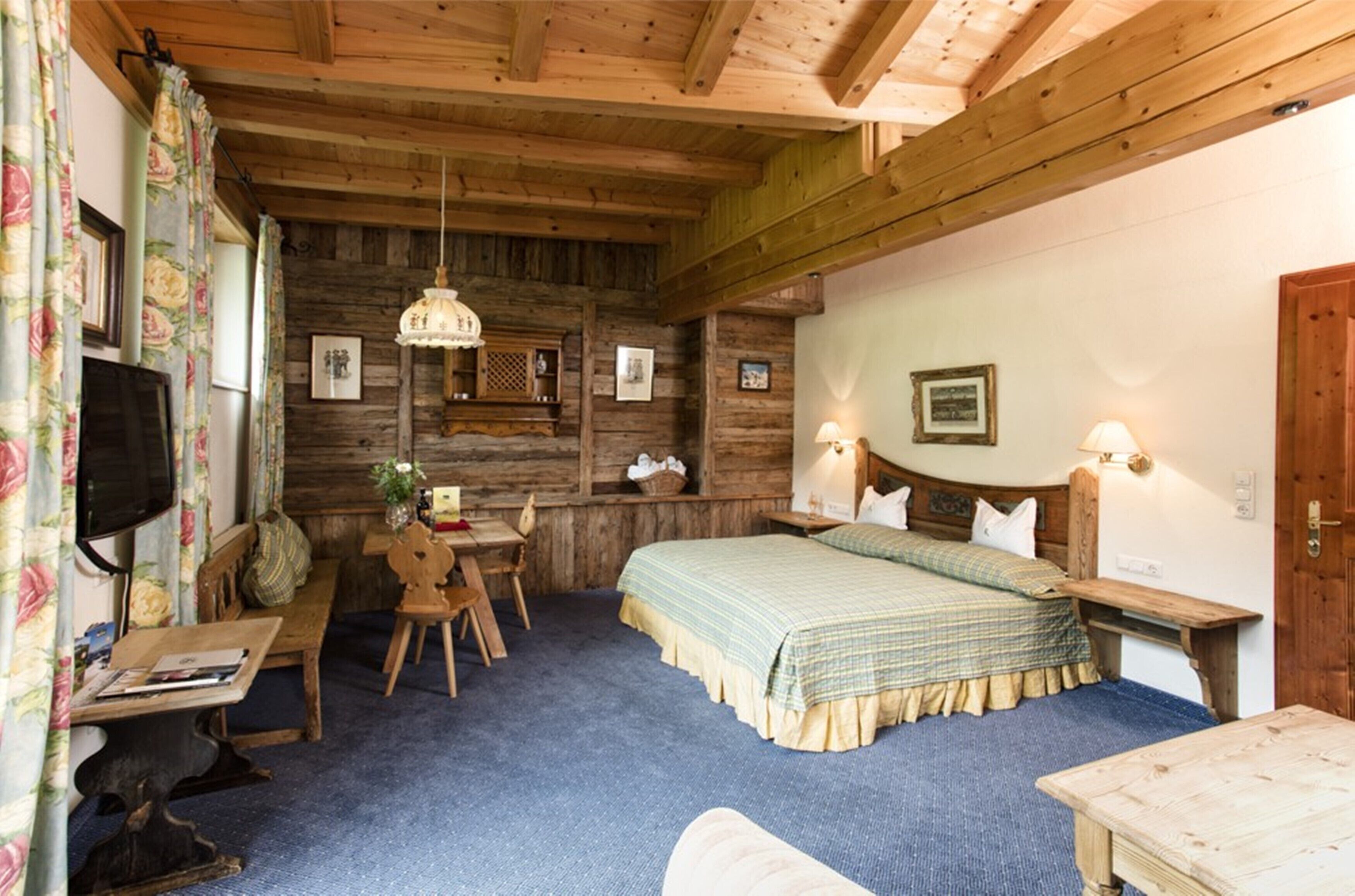 A rustic room with a wooden ceiling, a bed with a green checkered bedspread, a wooden desk, a chair, and a television on a stand.