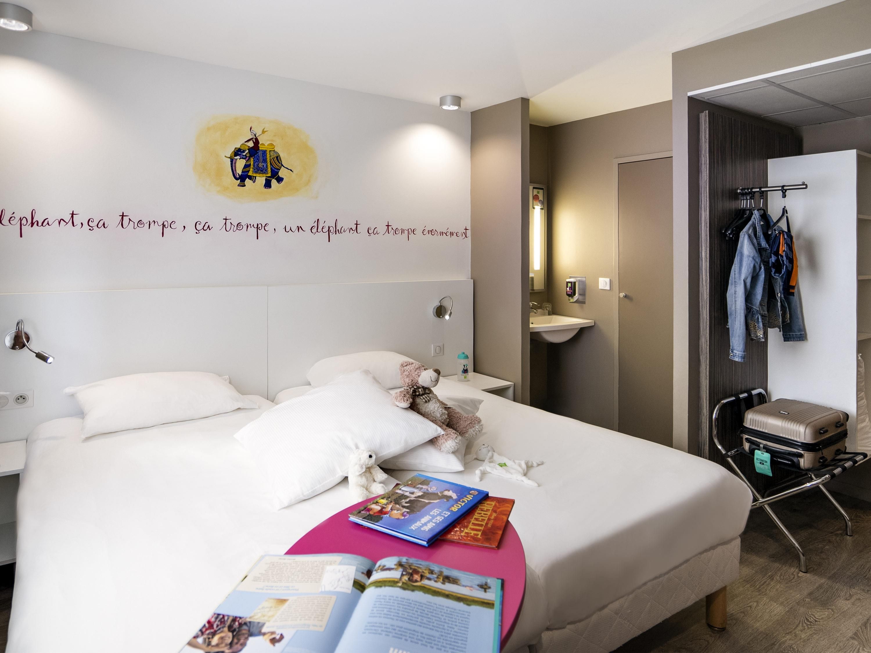 A hotel room with a large bed, a bedside table, a wall-mounted shelf, a hanging coat rack, and a suitcase on a trolley.