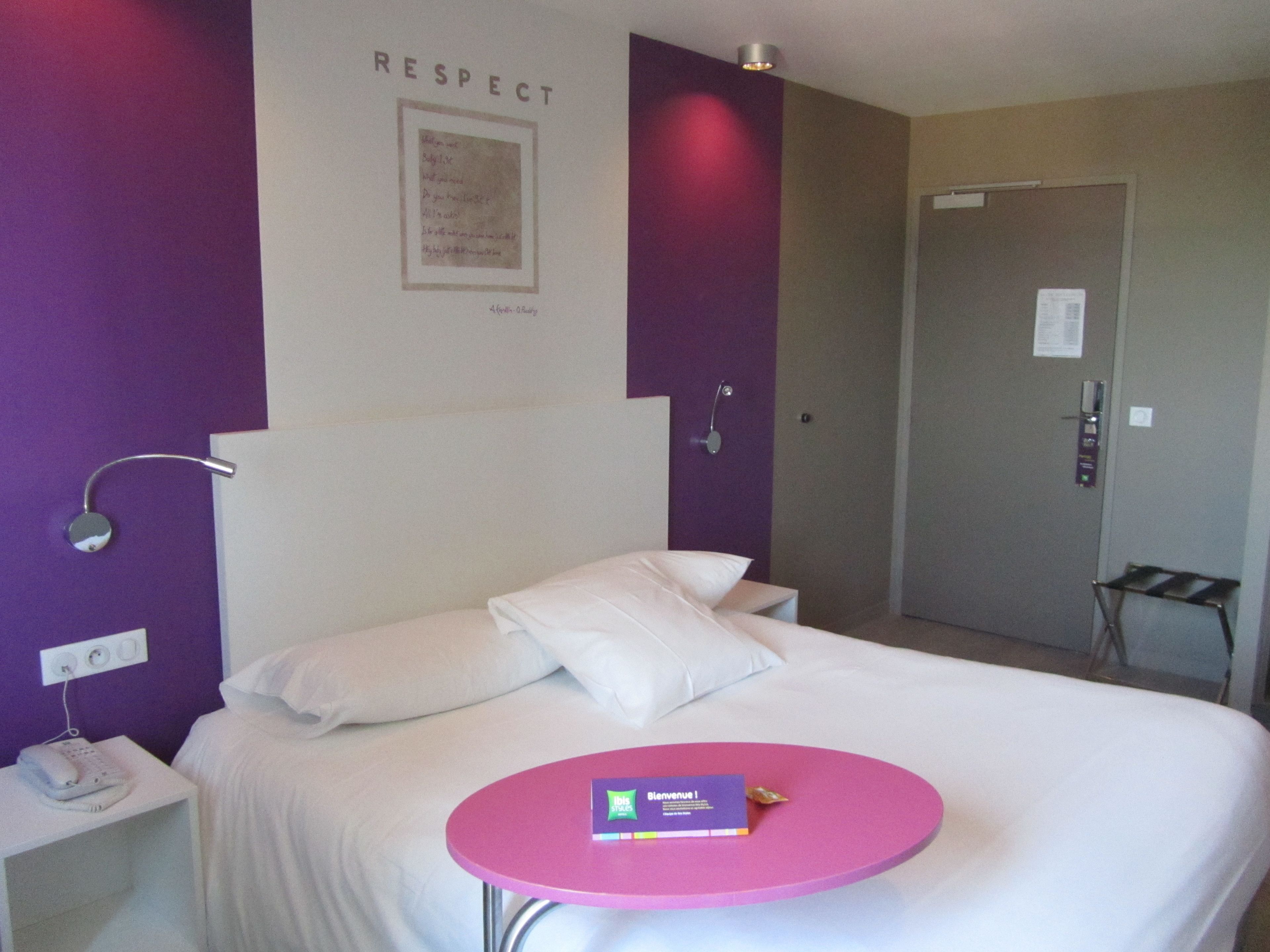 A hotel room with a bed, a bedside table with a phone, a wall-mounted lamp, and a door.