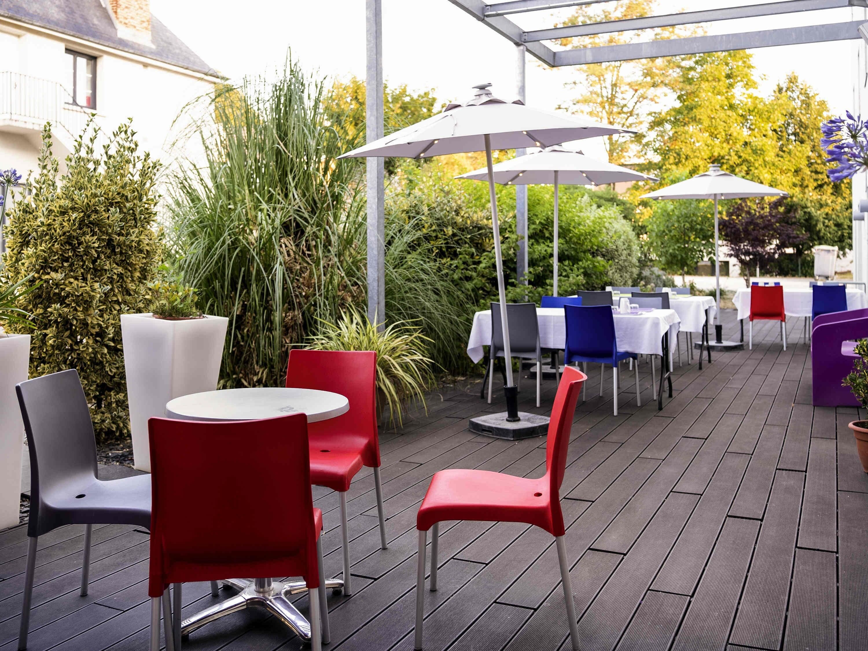 Ein Außenbereich mit einer Terrasse, runden Tischen und Stühlen, Sonnenschirmen und viel Grün.