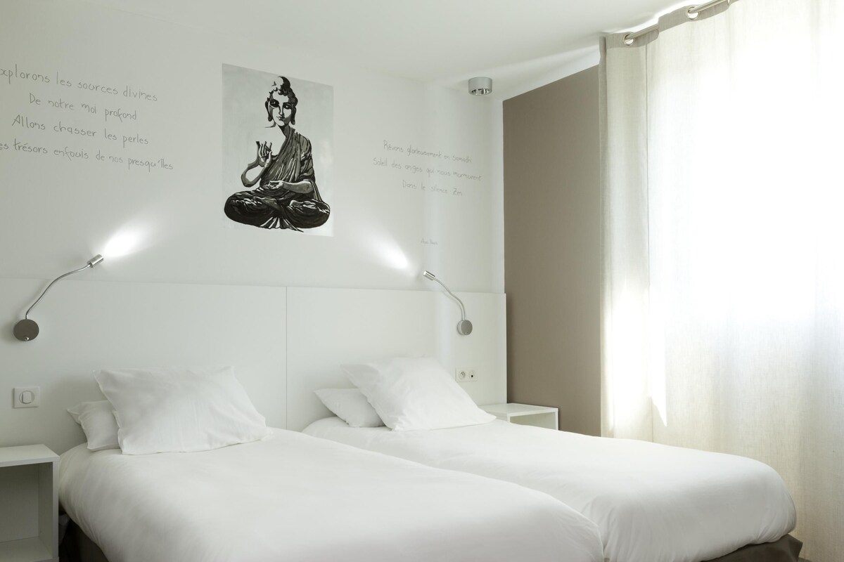 A hotel room with two beds, a wall-mounted light fixture, and a poster of a meditating figure.