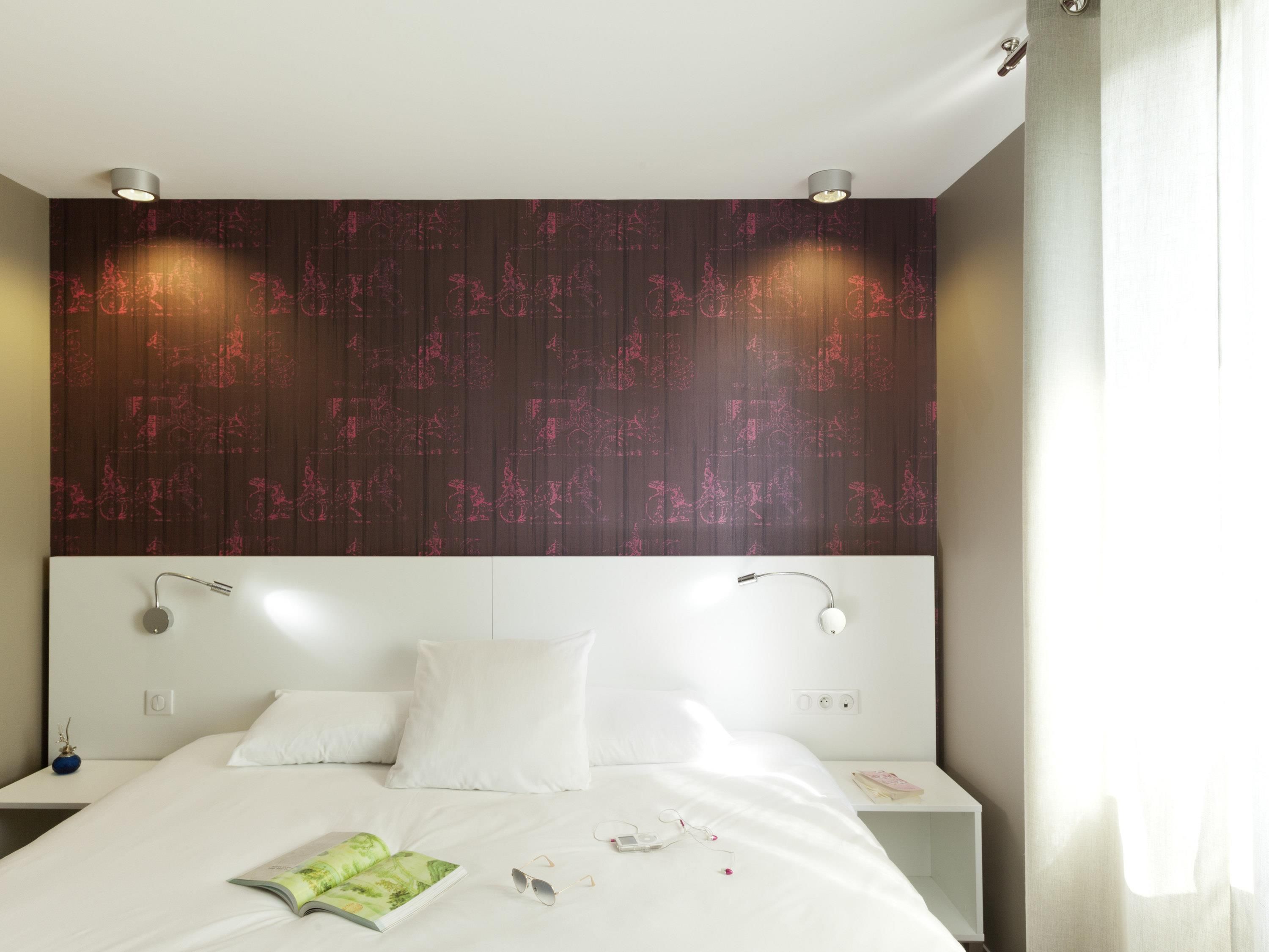 Una habitación de hotel moderna con una cama grande, ropa de cama blanca, cabecero con diseño estampado, mesitas de noche y una lámpara fijada a la pared.