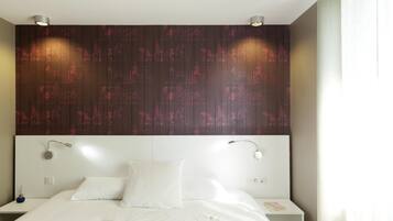 Una habitación de hotel moderna con una cama grande, ropa de cama blanca, cabecero con diseño estampado, mesitas de noche y una lámpara fijada a la pared.