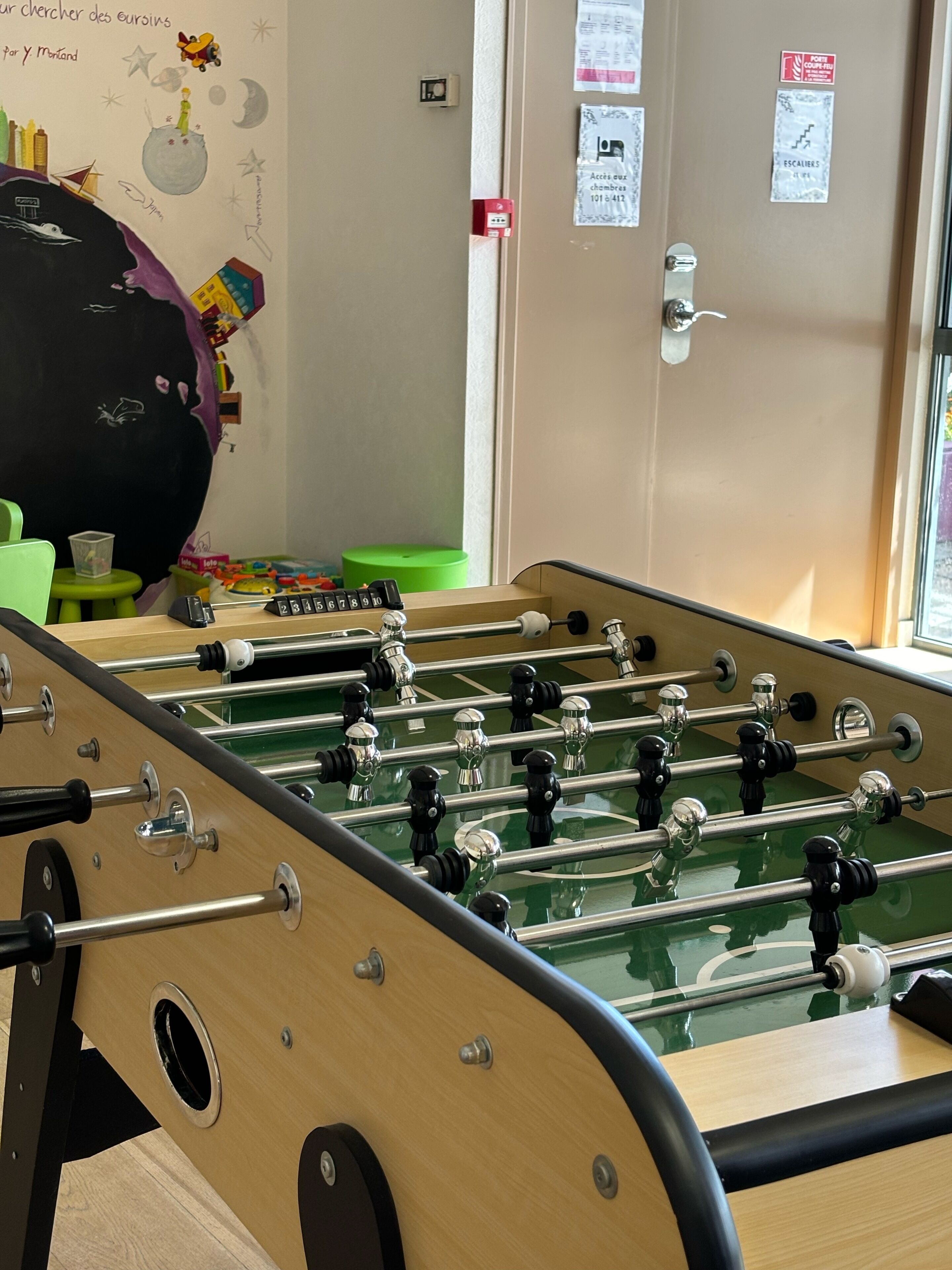 A foosball table in a room with a wall mural, a green chair, and a door with posted notices.