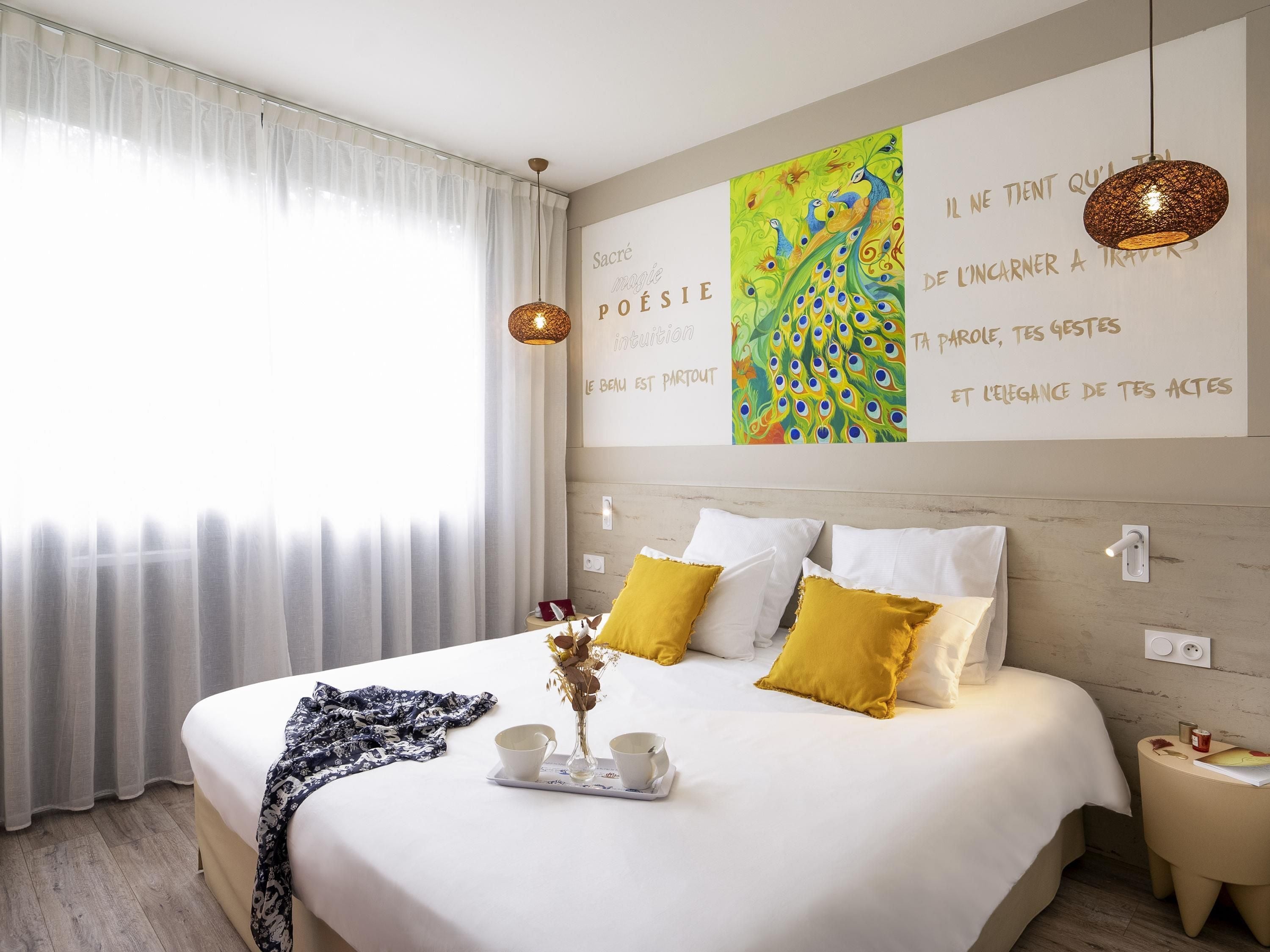 A neatly made bed with white and mustard pillows, a vase with flowers, and a tray with cups.