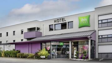 Un hotel de diseño moderno, con un llamativo letrero de 'ibis Styles', un acceso cubierto y un cartel que indica la disponibilidad de estacionamiento.