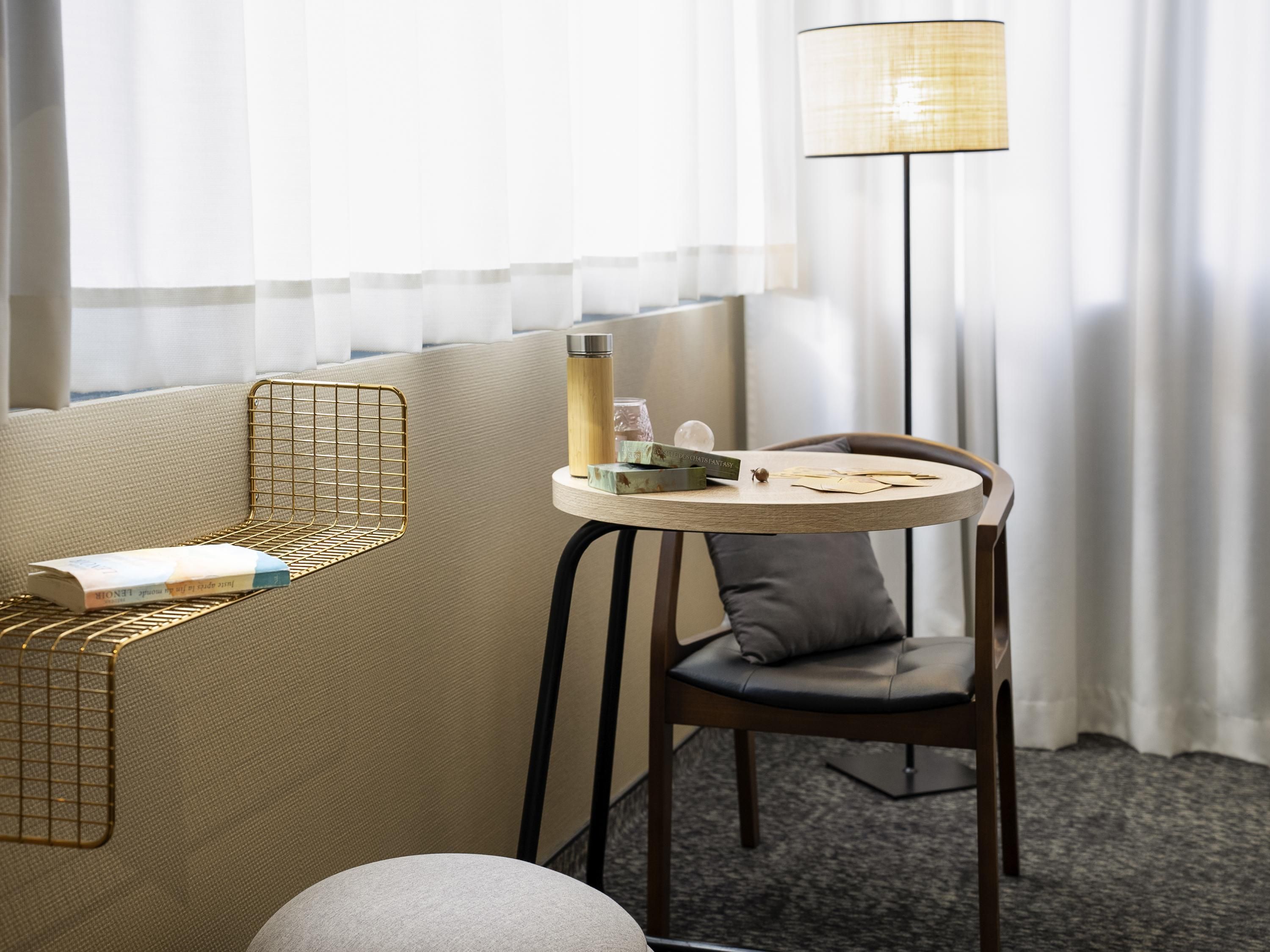 A hotel room with a round table, a chair, a floor lamp, a bookshelf, and a window with sheer curtains.