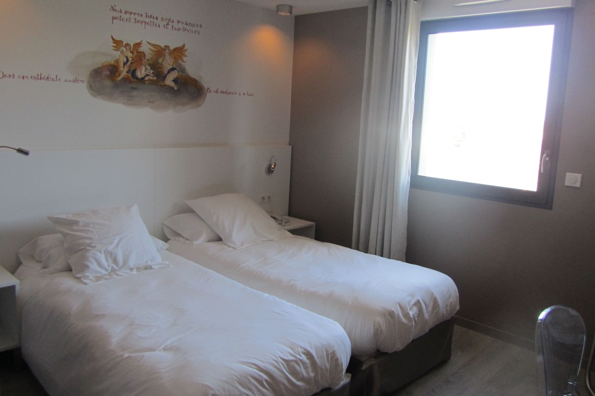 Una camera d'albergo con due letti singoli, un comodino con una lampada, una finestra con tende e un pannello decorativo al muro.