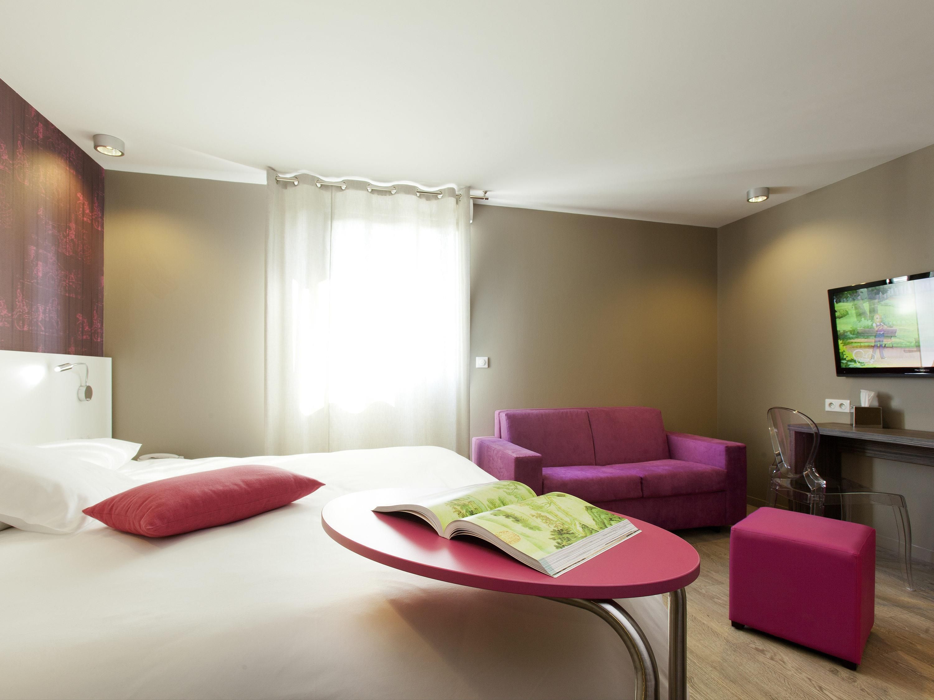 Una habitación de hotel con una cama, una mesita con una revista, un sillón morado, un reposapiés rosa y un televisor en la pared.