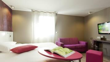 Una habitación de hotel con una cama, una mesita con una revista, un sillón morado, un reposapiés rosa y un televisor en la pared.