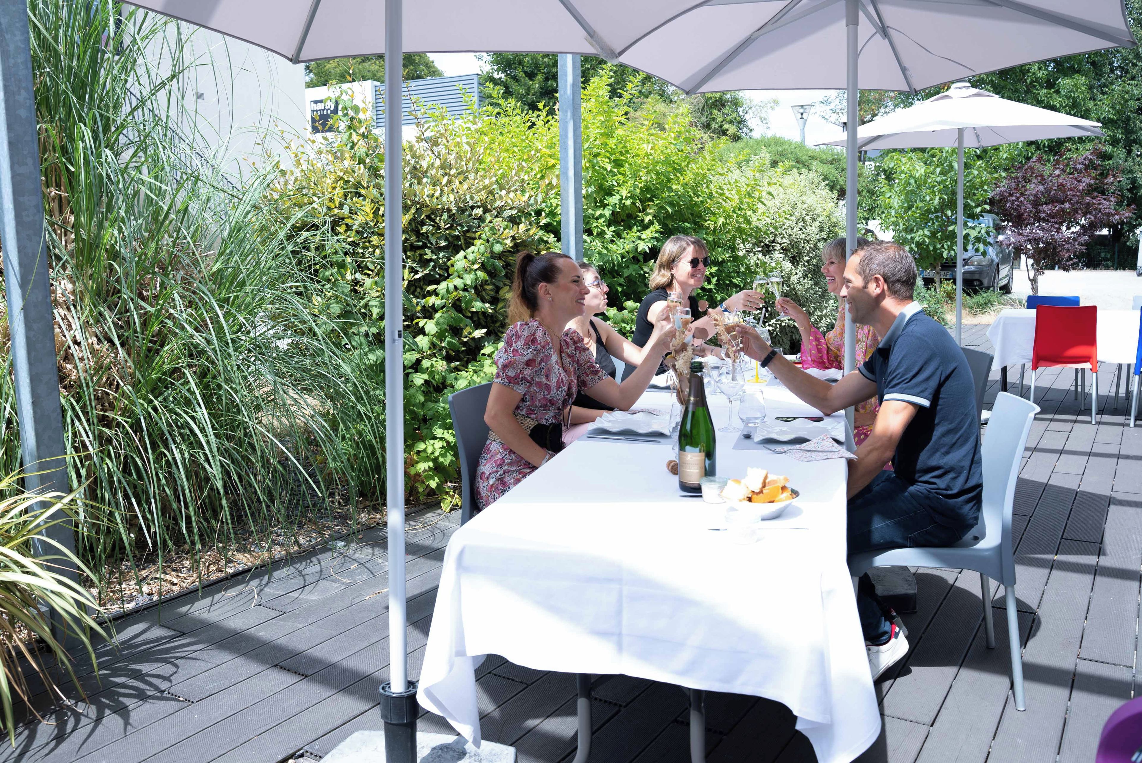 A group of people seated at an outdoor table with a white tablecloth, surrounded by greenery and umbrellas.