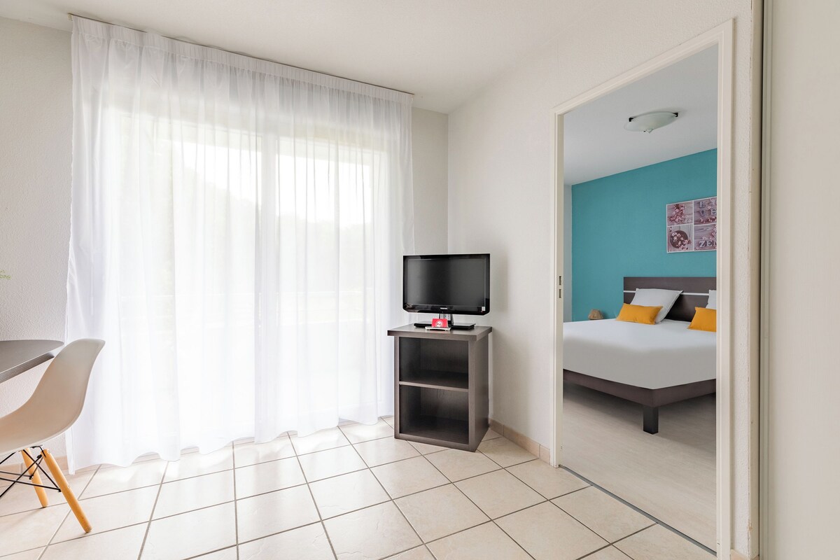 A hotel room with a bed, a TV on a stand, a desk with a chair, and a window with sheer curtains.