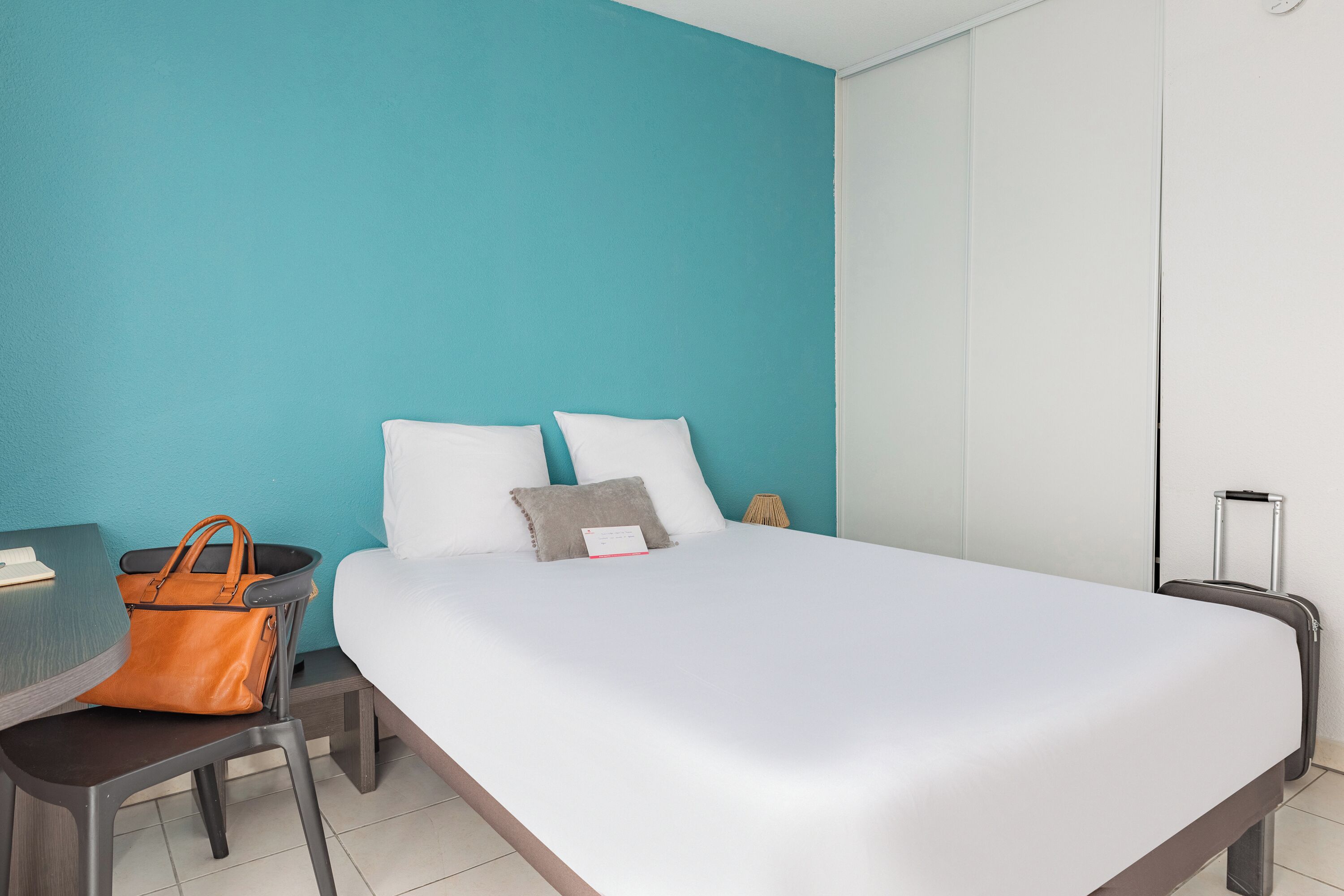 Habitación de hotel con cama, escritorio, silla, maleta y pared color verde azulado.
