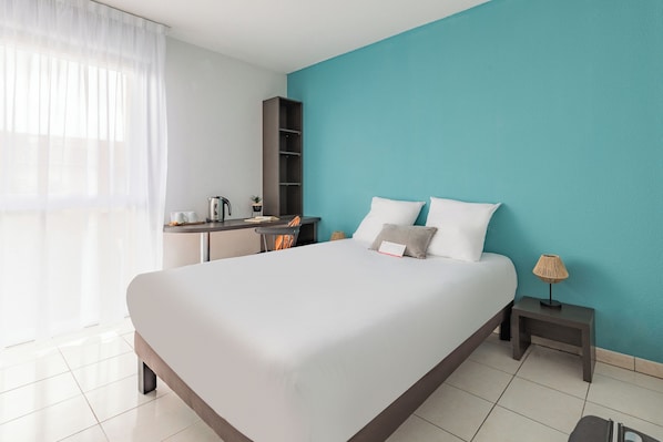 Ein modernes Hotelzimmer mit einem großen Bett, einem Schreibtisch mit Stuhl, einem kleinen Tisch mit Lampe und einem Kleiderschrank.