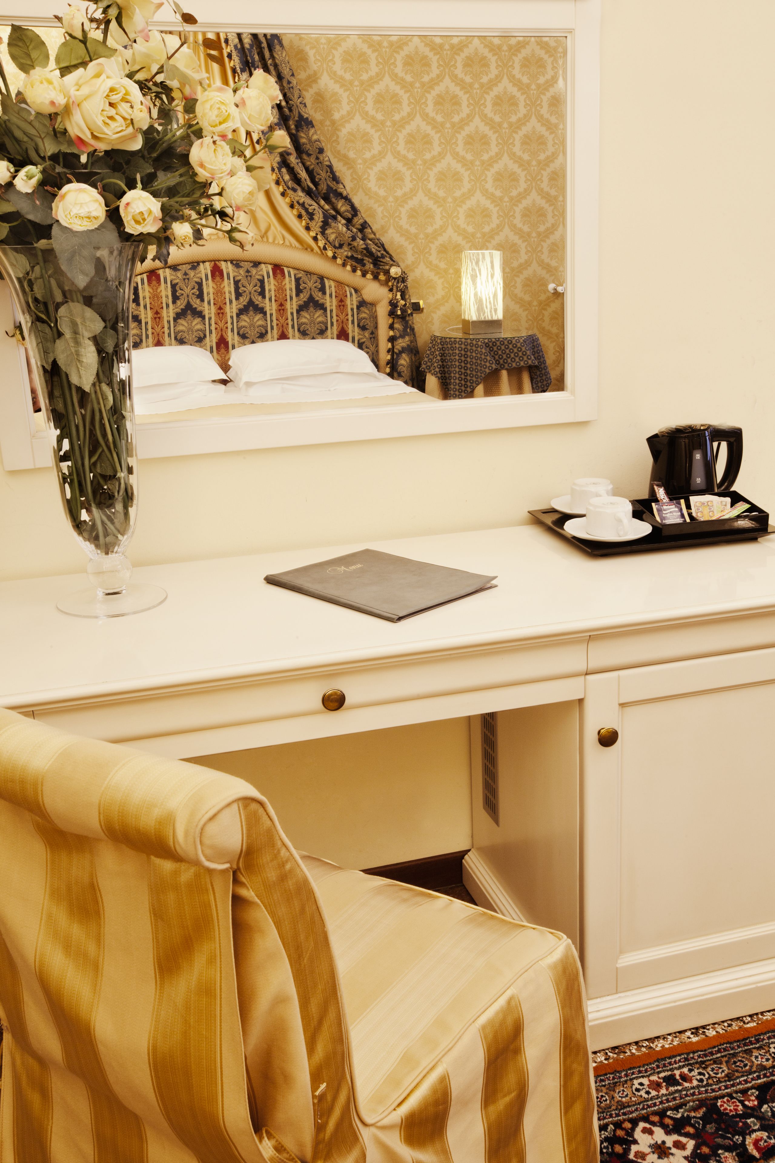 Habitación de hotel con escritorio blanco, un jarrón de flores, un espejo, mesita de noche con lámpara y cafetera.