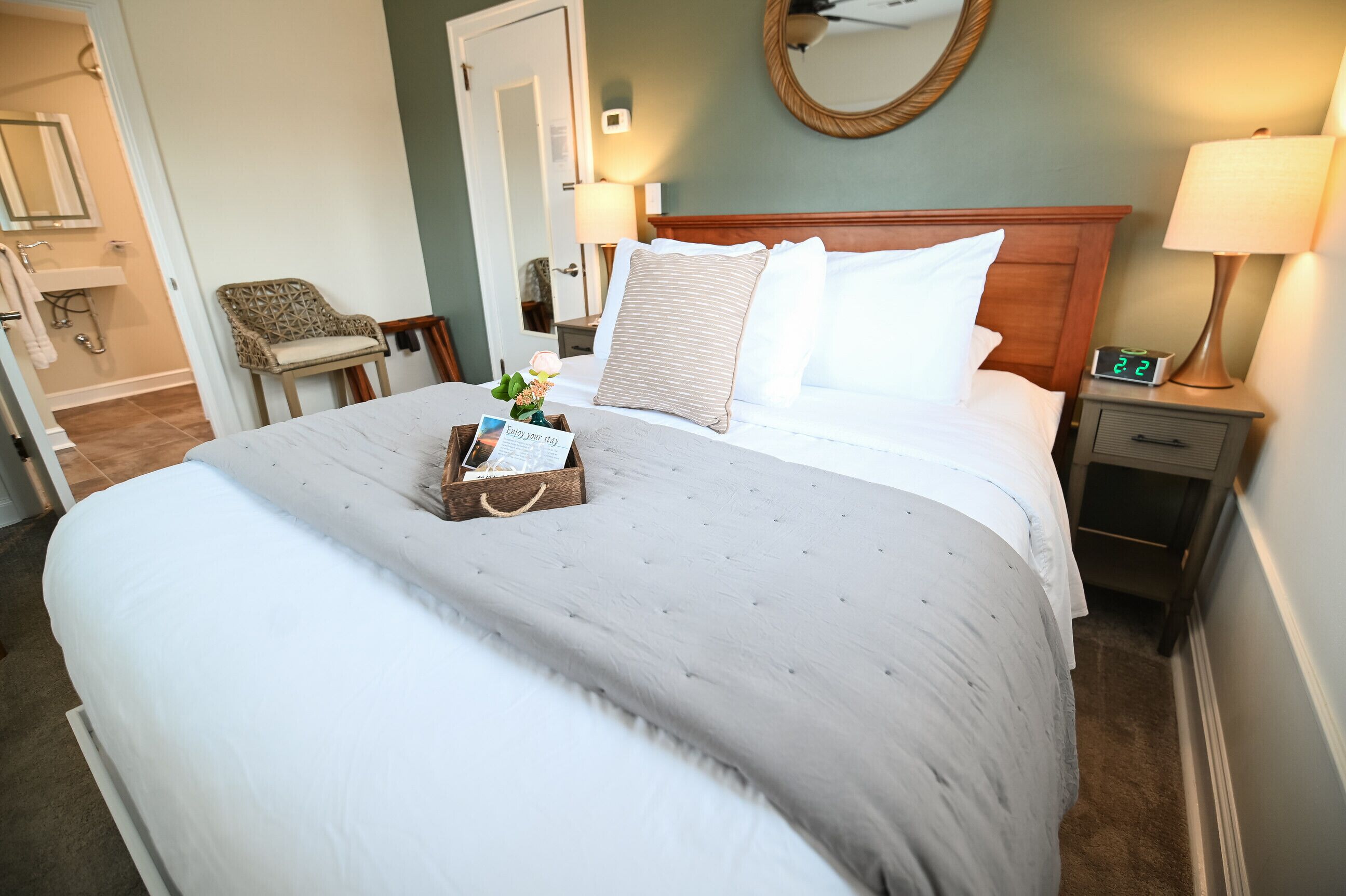 A neatly made bed with a grey bedspread, a wooden nightstand with a lamp, and a chair in a hotel room.
