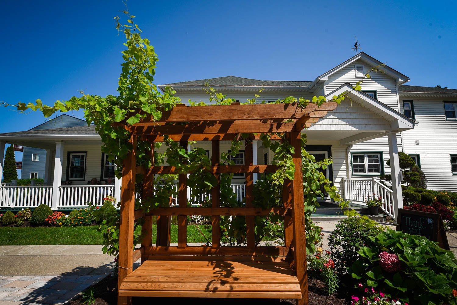 A wooden pergola with vines, a bench, and a garden in front of a two-story house with a porch.