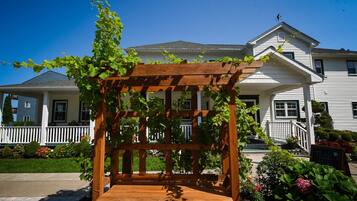 A wooden pergola with vines, a bench, and a garden in front of a two-story house with a porch.