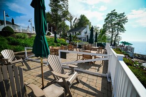 Une terrasse dotée de fauteuils Adirondack et de parasols, offrant une vue sur un plan d’eau et une maison blanche.
