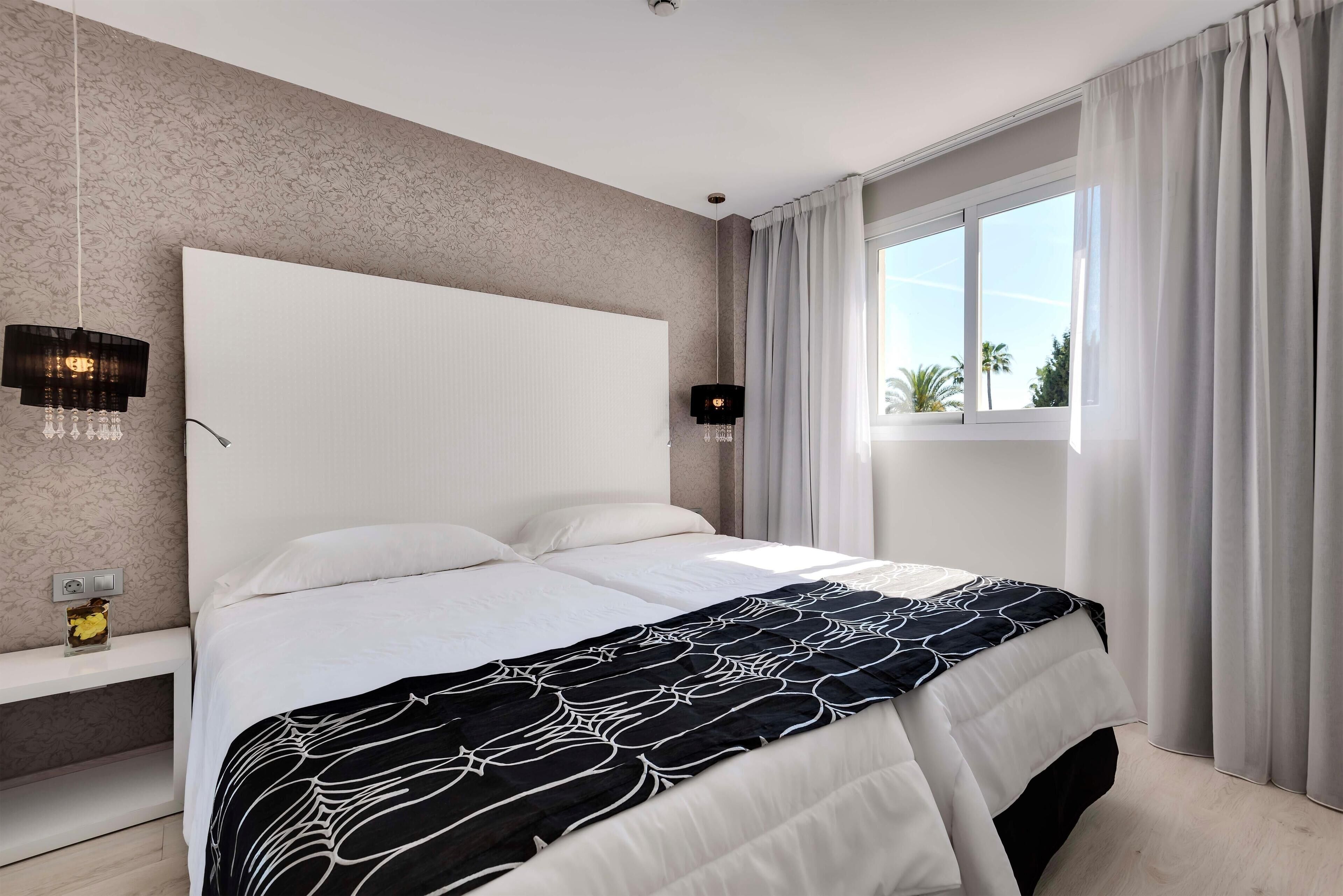 Una cama bien hecha con una colcha estampada, una mesita de noche con lámpara y una ventana con cortinas.