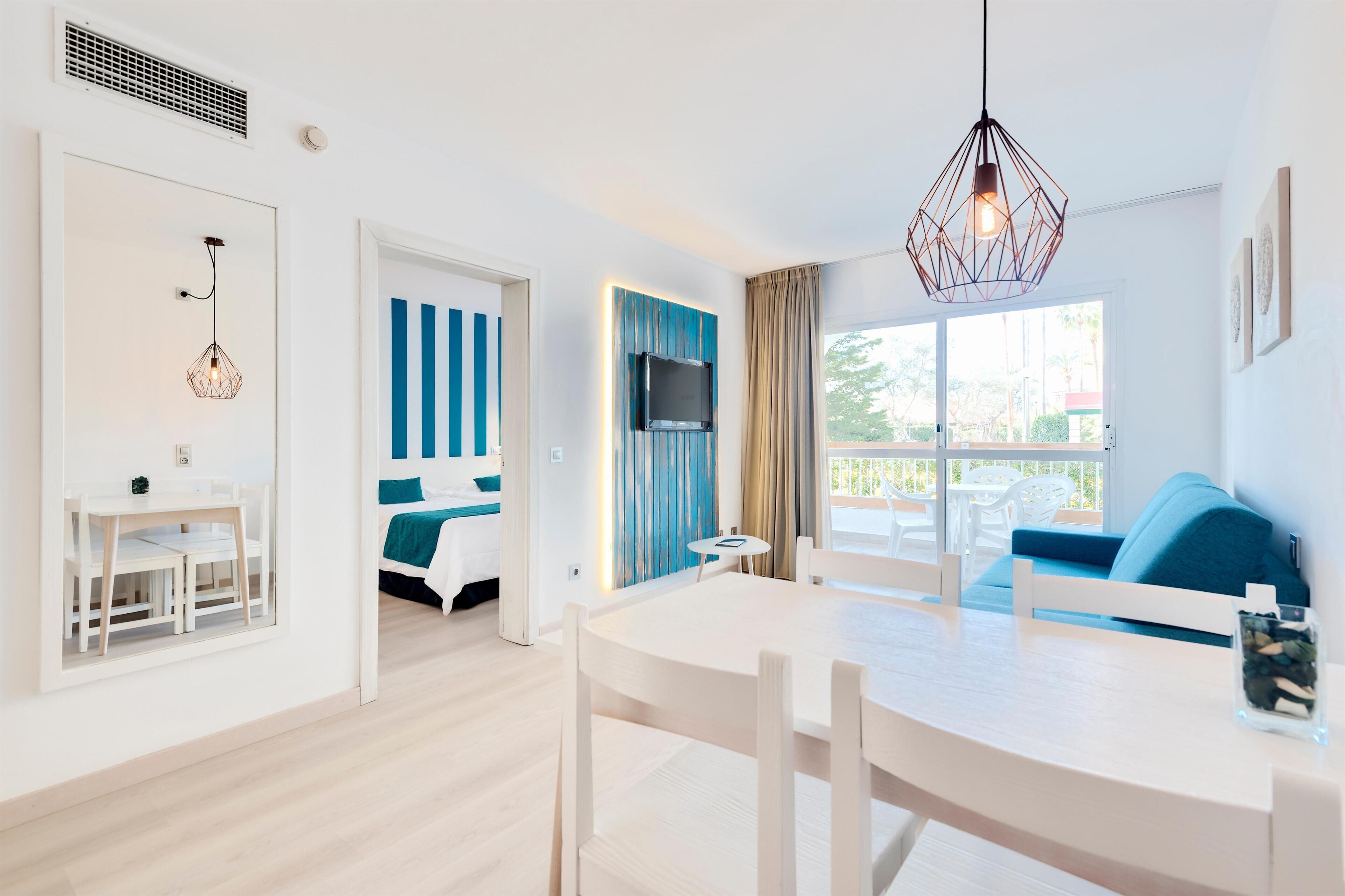 Habitación de hotel moderna con zona de comedor, televisor montado en la pared y balcón con vistas.