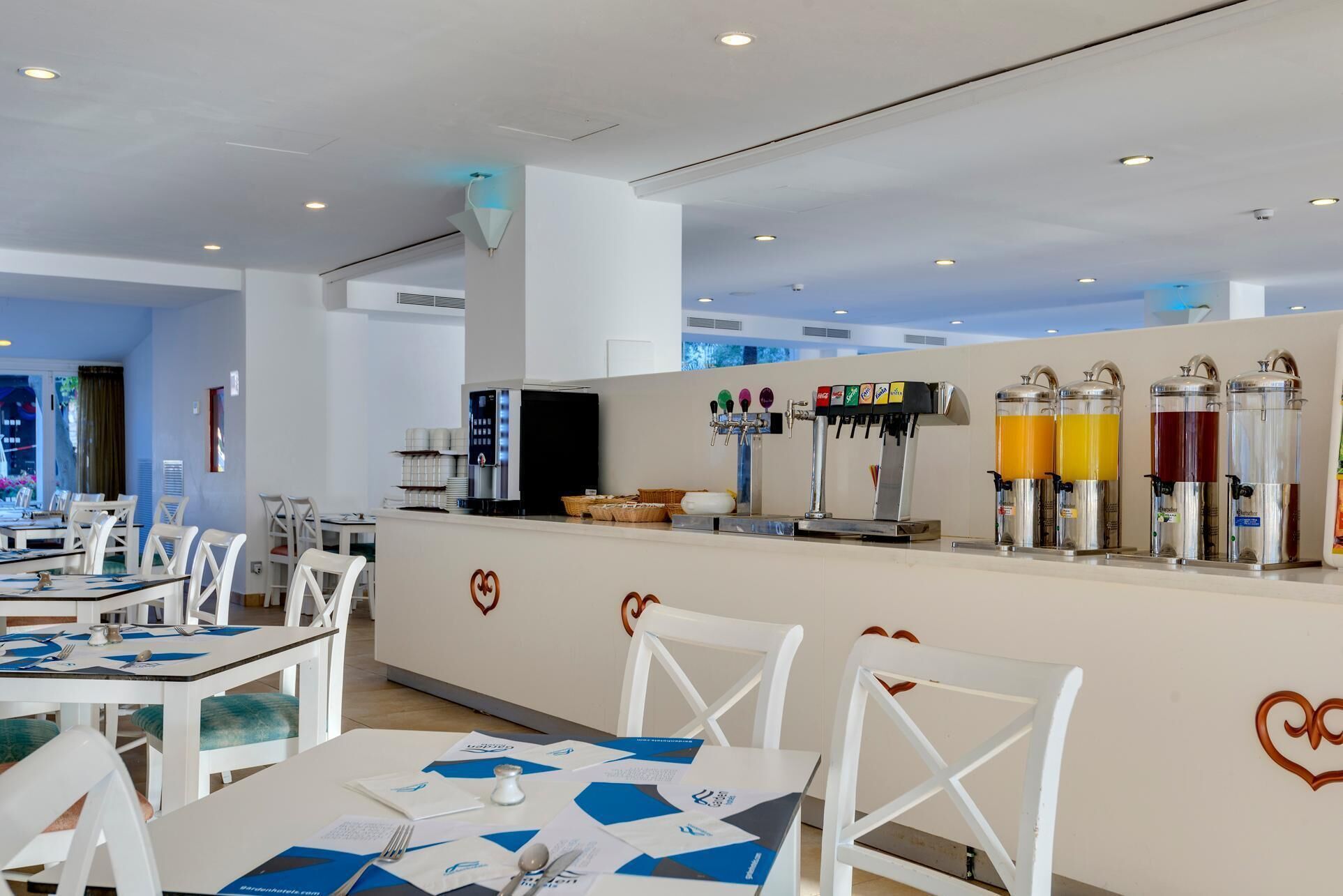 Comedor de hotel moderno con mesas y sillas blancas, una estación de bebidas de autoservicio y un logotipo con forma de corazón en la pared.